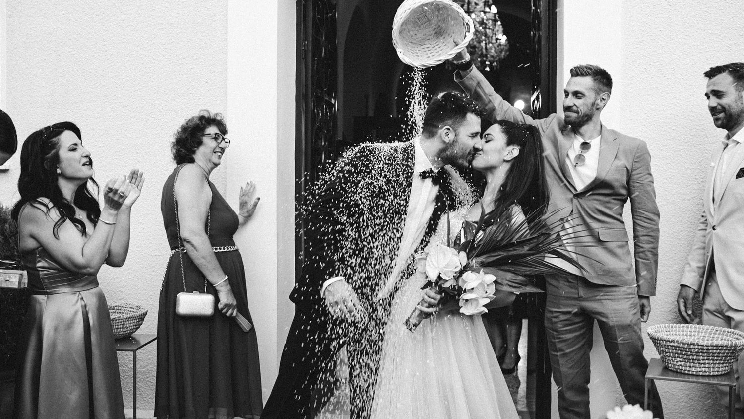 A black and white photo of wedding guests celebrating and cheering as a couple shares a festive moment at their reception.