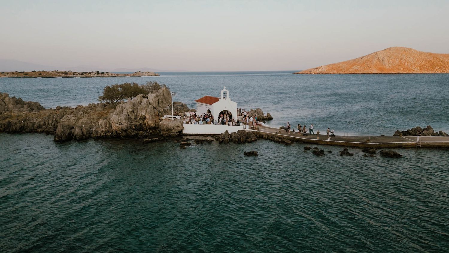 A small white church stands on rocky outcrop surrounded by turquoise Mediterranean waters at sunset.