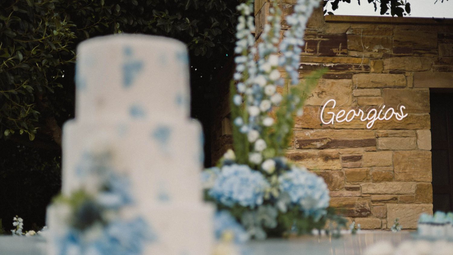 A blue and white wedding cake blurred in foreground with stone wall and blue hydrangeas in rustic wedding setting.