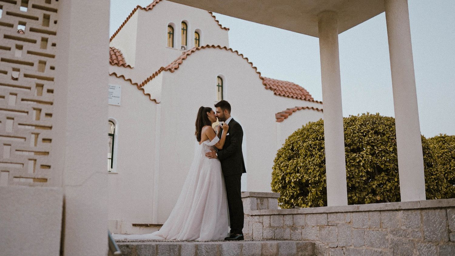 Newlywed couple embraces under a white columned portico of a Spanish mission-style church with adobe architecture.
