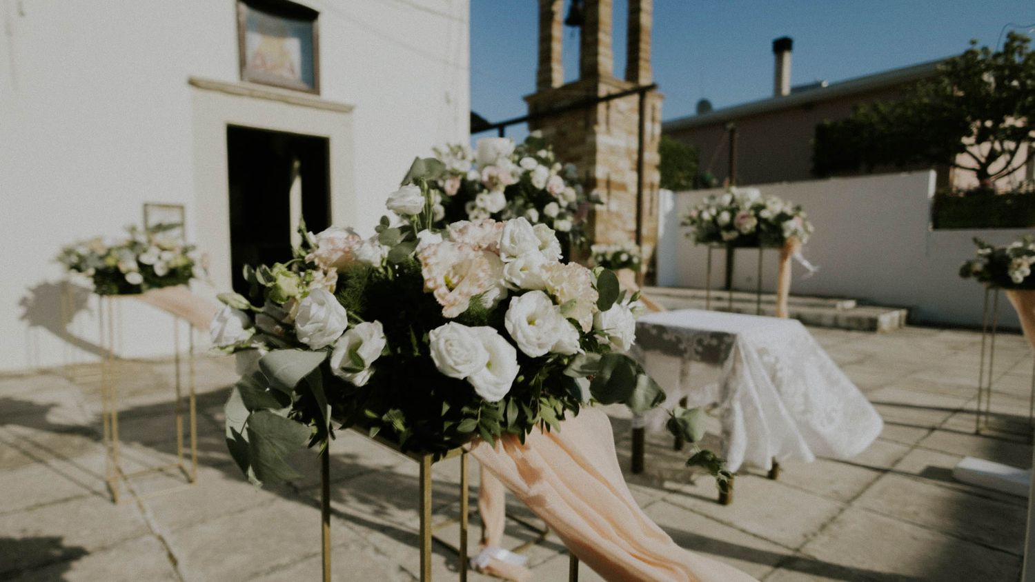 Elegant white floral arrangements line a sunlit outdoor wedding ceremony space with stone architecture in the background.