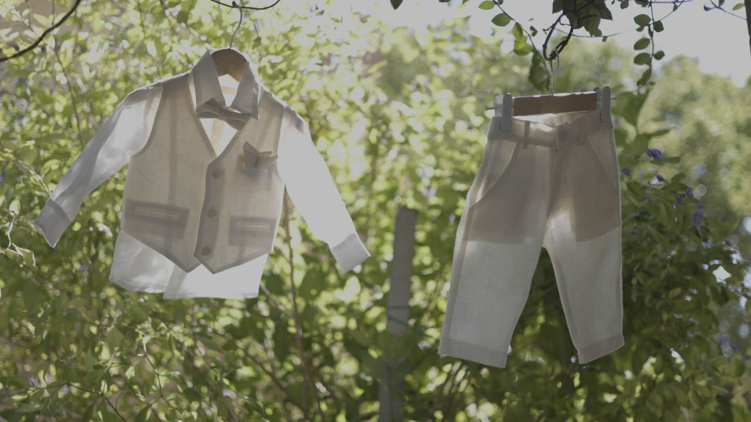 Baby clothing drying on a clothesline outdoors with a white dress shirt and gray pants against green foliage background.