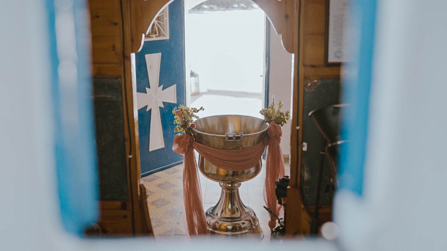 A golden baptismal font viewed through a church doorway with a cross visible in the background.