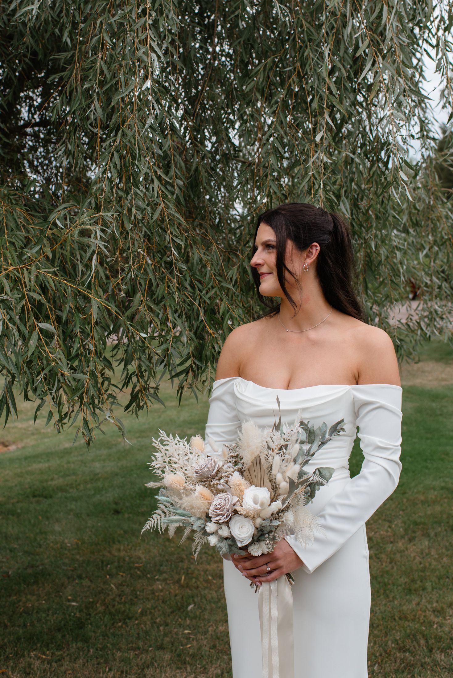 An elegant off-shoulder white wedding dress with long sleeves complemented by a dried floral bouquet under a weeping willow tree.