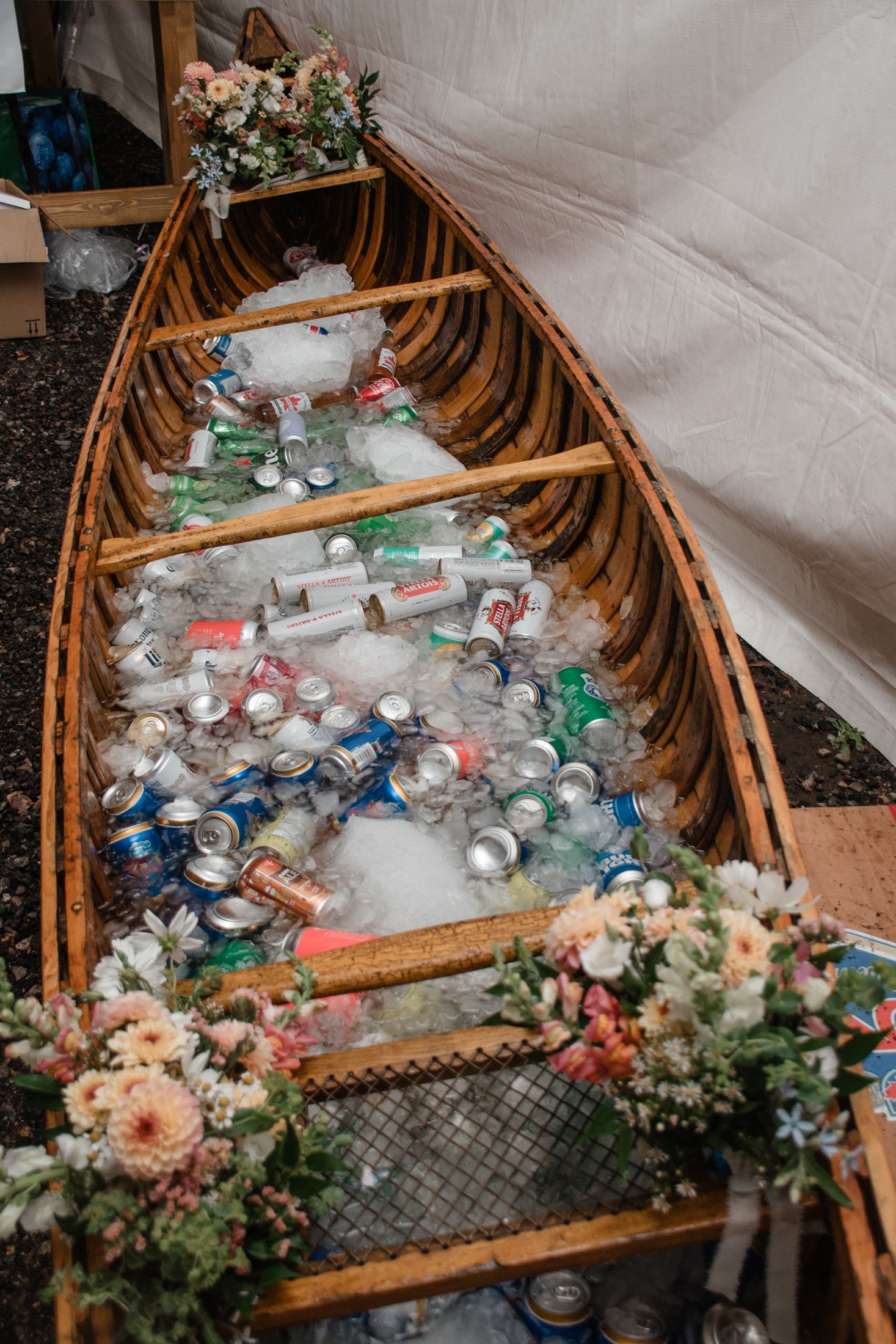 Vintage wooden canoe filled with ice and beverages decorated with floral arrangements at a rustic outdoor event.