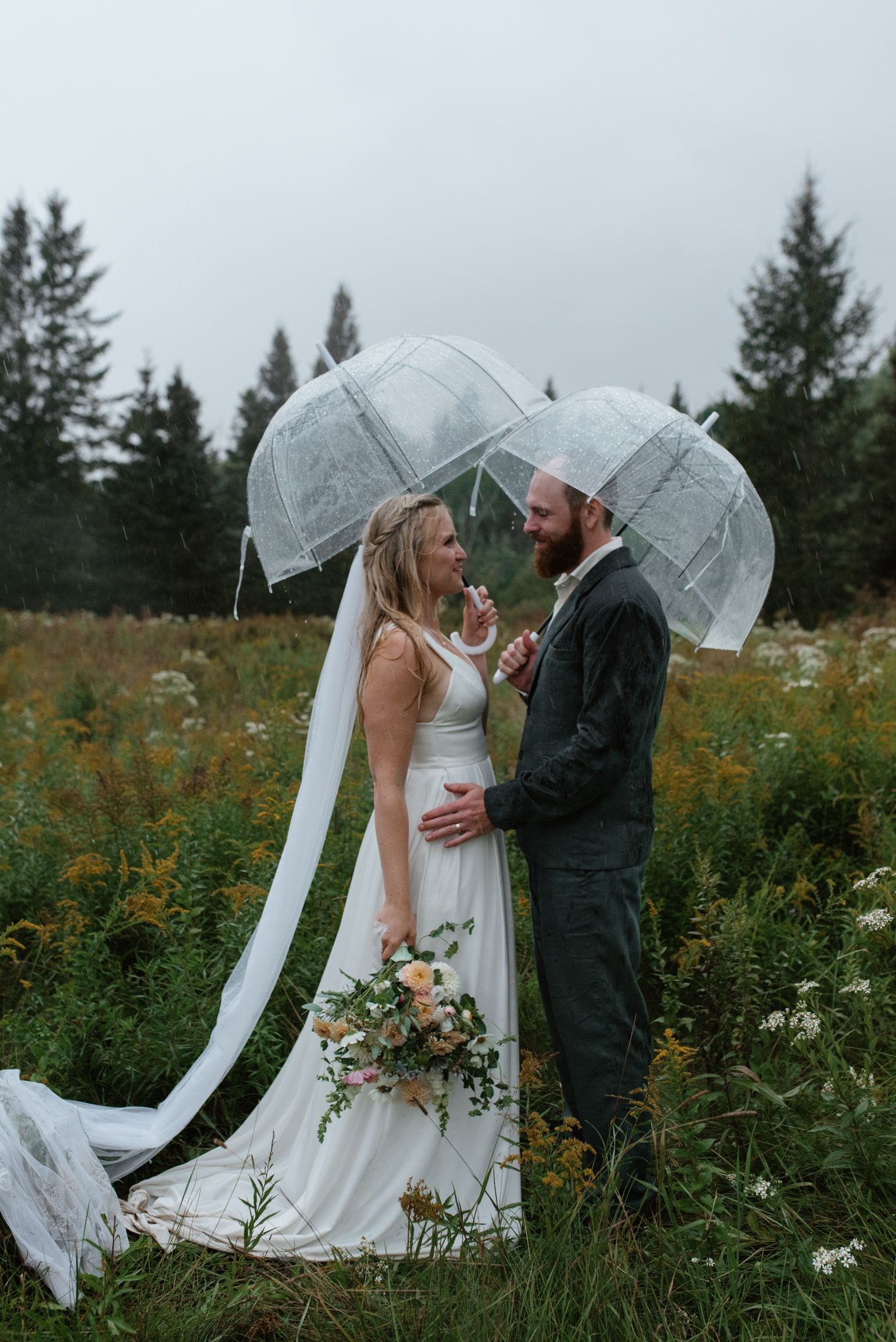 A couple share an intimate moment under clear umbrellas in a misty meadow with evergreen trees in the background.