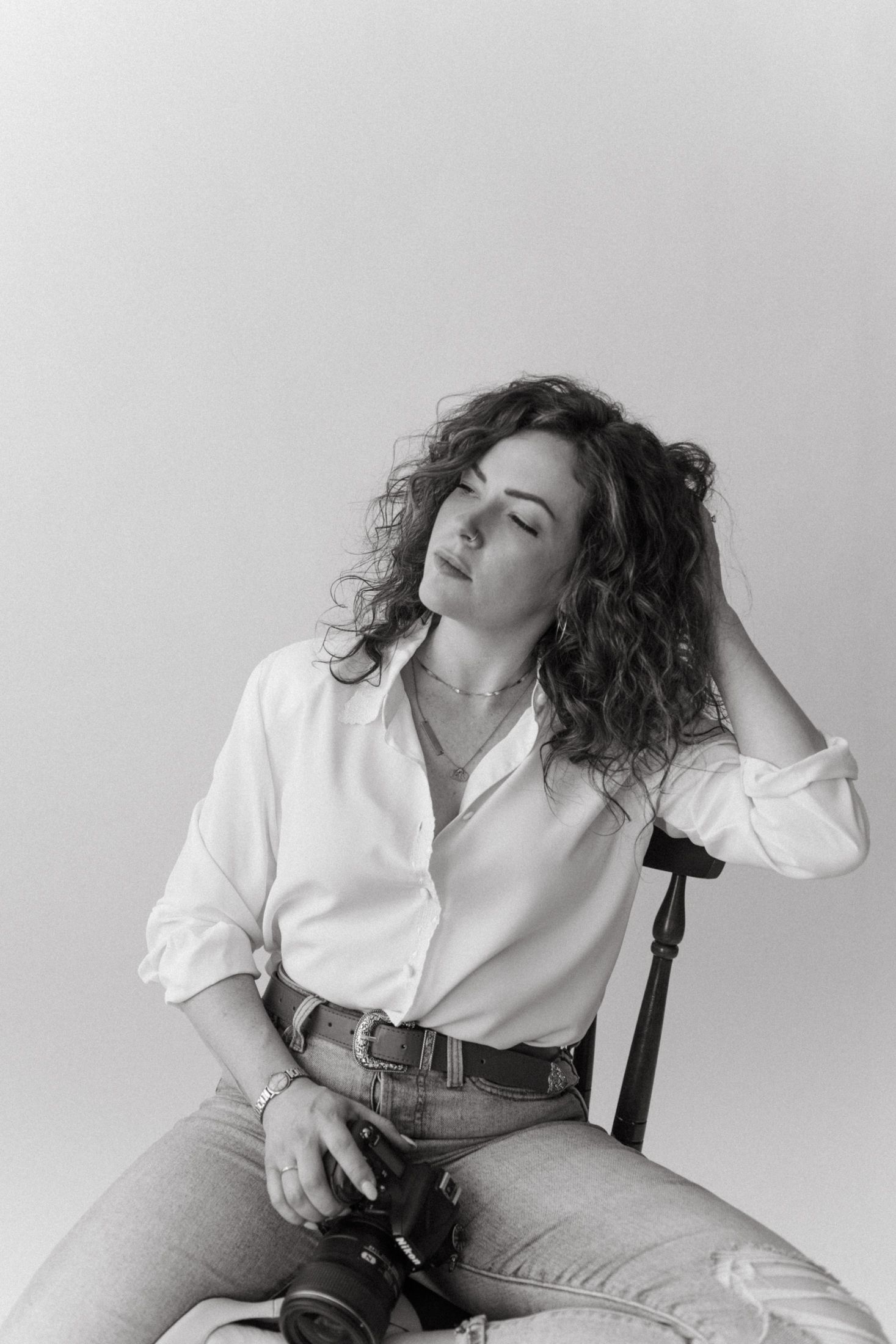 A figure in a white button-down shirt and jeans reclines dramatically in a chair against a light background for a fashion photo.