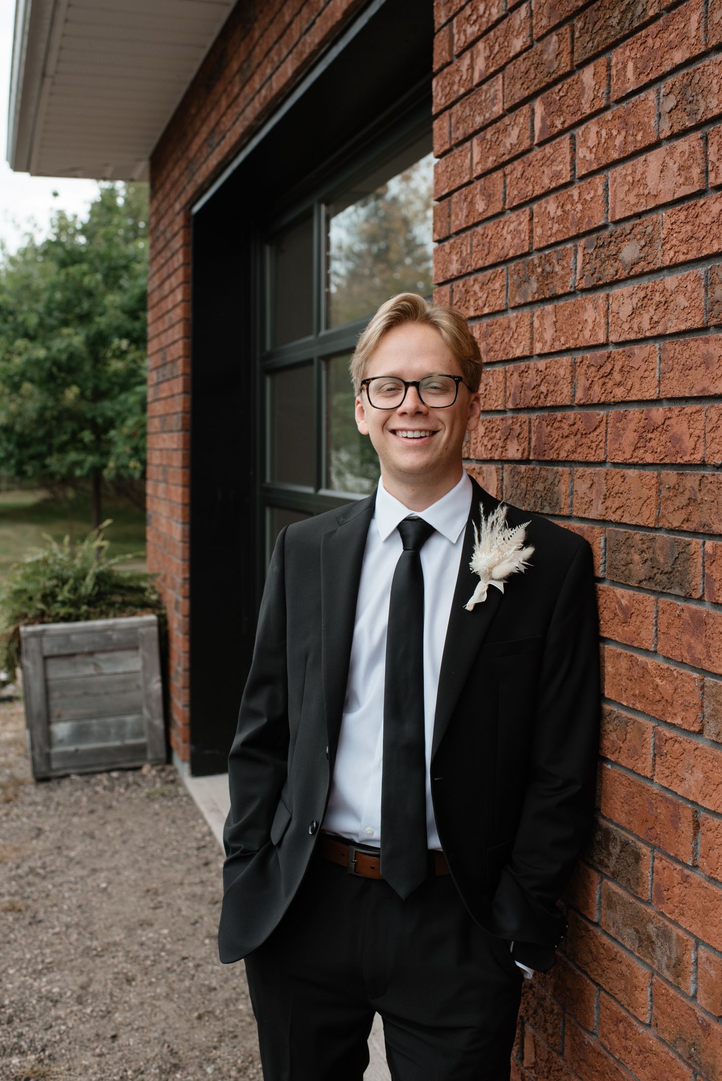 A groom in a classic black suit and glasses leans against a brick wall with a boutonniere on his lapel.