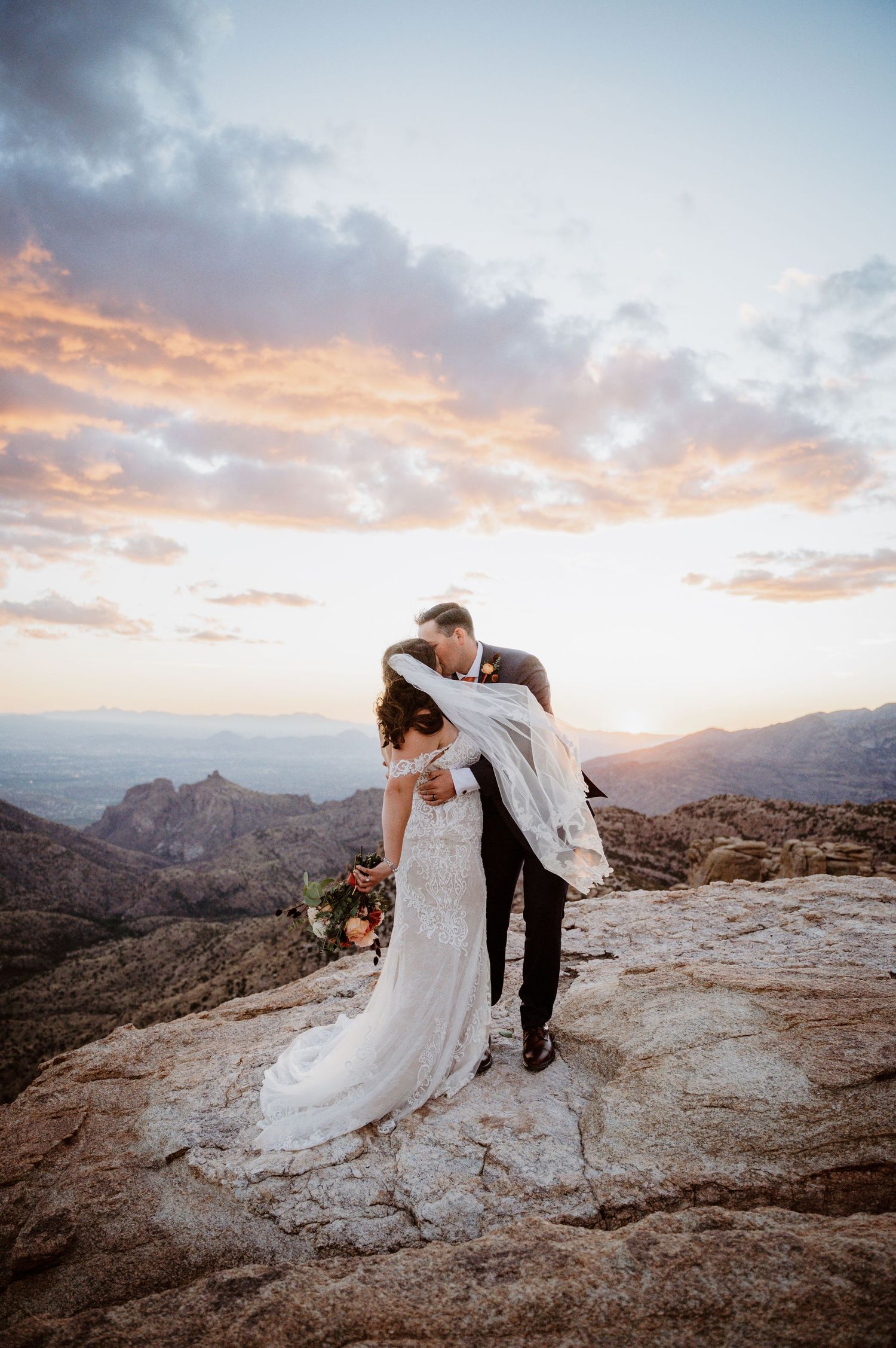 A romantic wedding couple embraces on a mountain peak at sunset with dramatic clouds and desert landscape views.