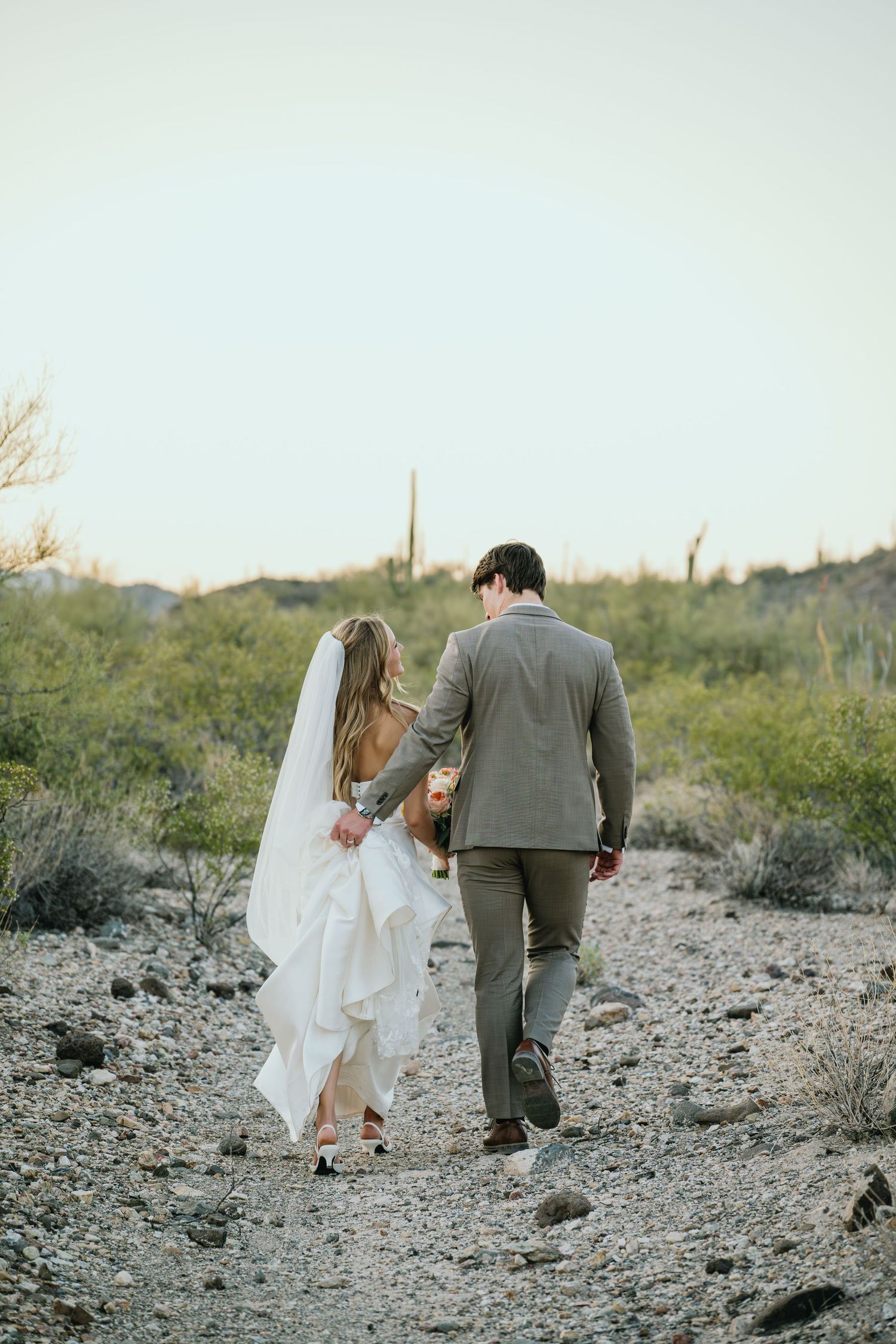A couple in wedding attire walking through a desert landscape with saguaro cacti in the background at sunset.