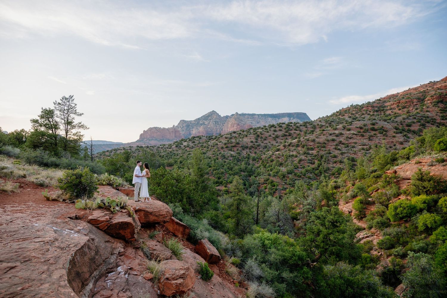 Scenic view of red rock cliffs and desert landscape in Sedona with mountains and vegetation under cloudy skies.