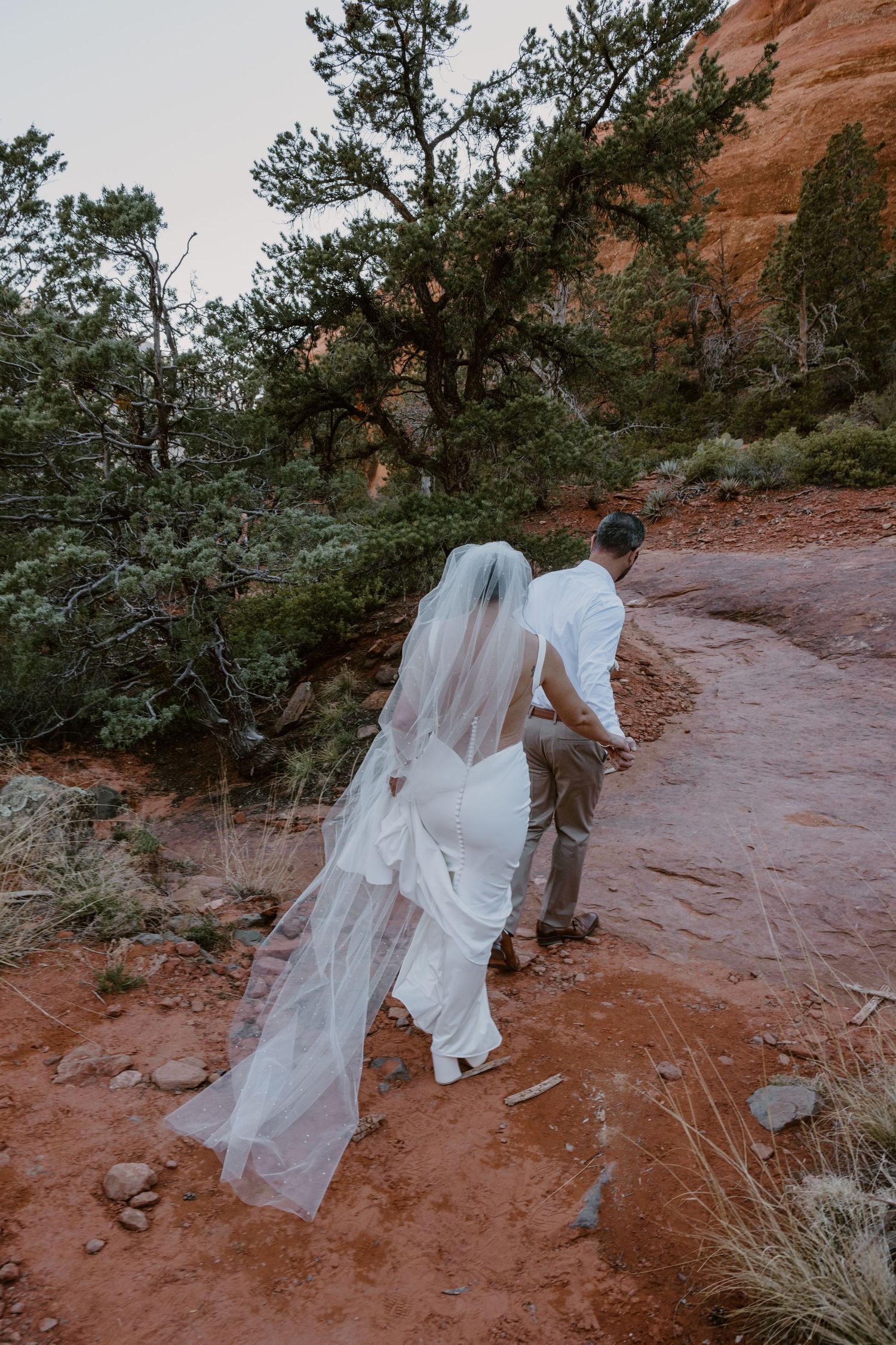 A couple in wedding attire walk along a winding red rock trail surrounded by desert vegetation and sandstone cliffs.