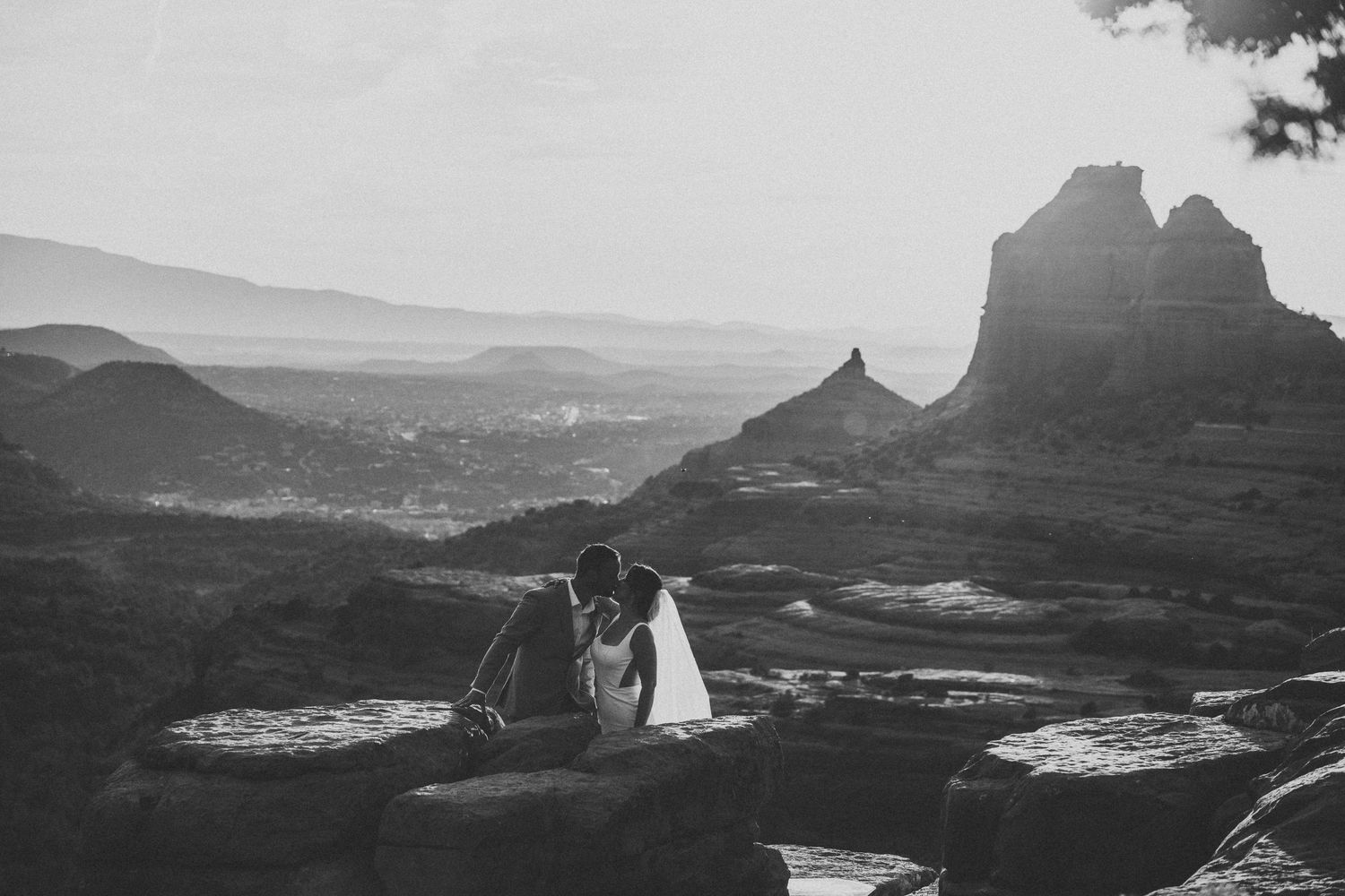A couple sits together on red rocks overlooking the dramatic landscape of Sedona Arizona at sunset in black and white.
