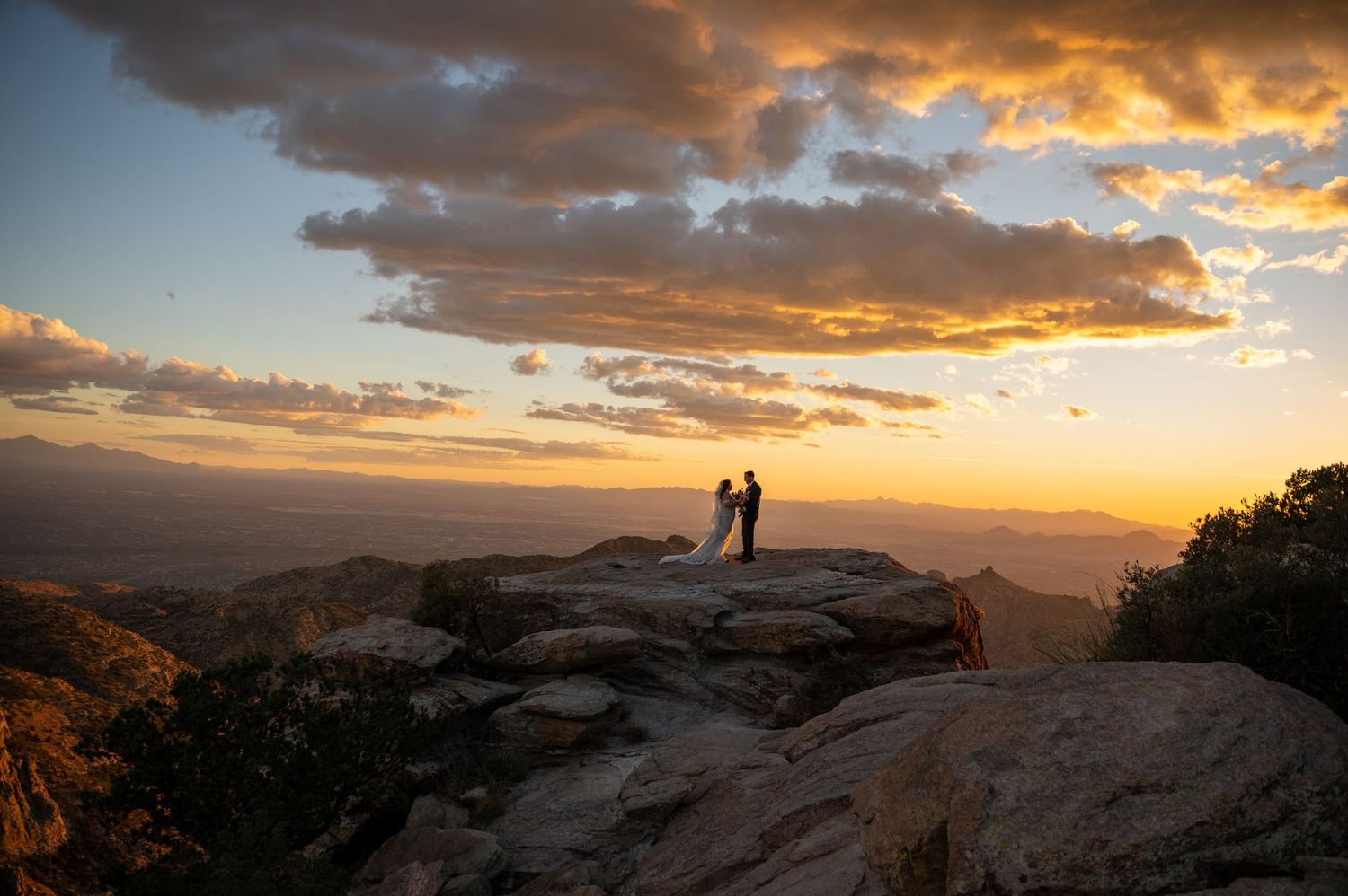 A silhouetted figure stands atop a rocky mountain peak during a dramatic golden sunset with clouds streaking the sky.