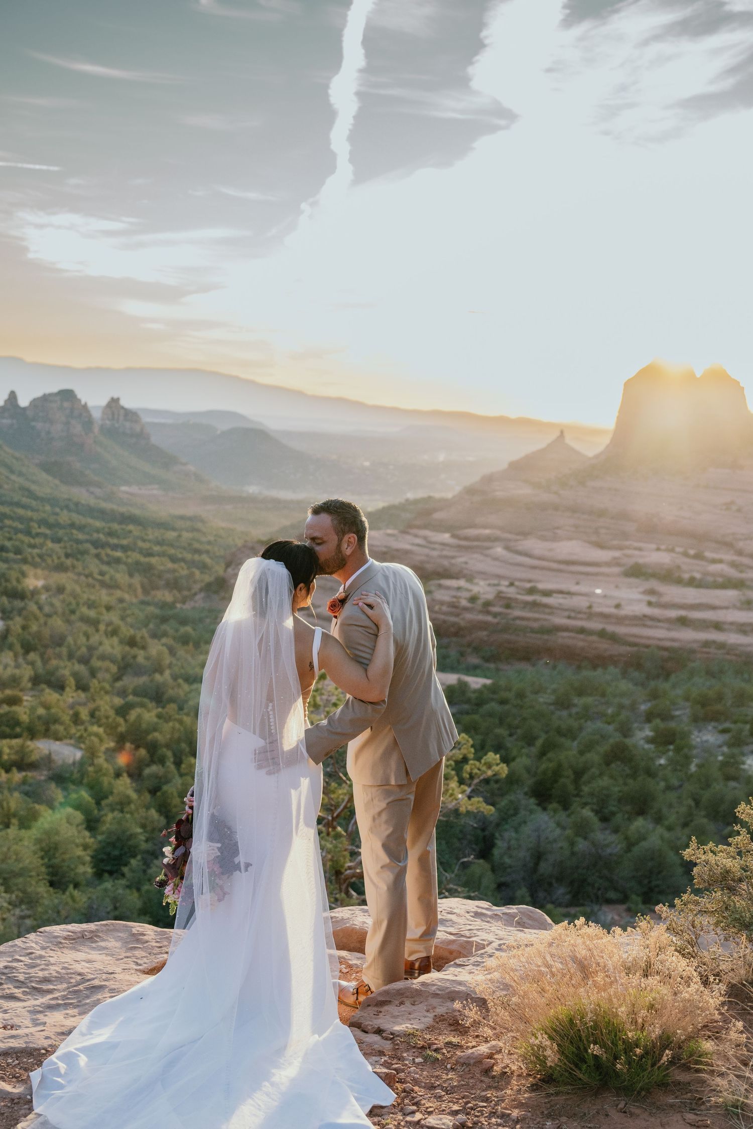 A couple shares their first kiss as newlyweds against a stunning backdrop of red rock formations in Sedona at sunset.