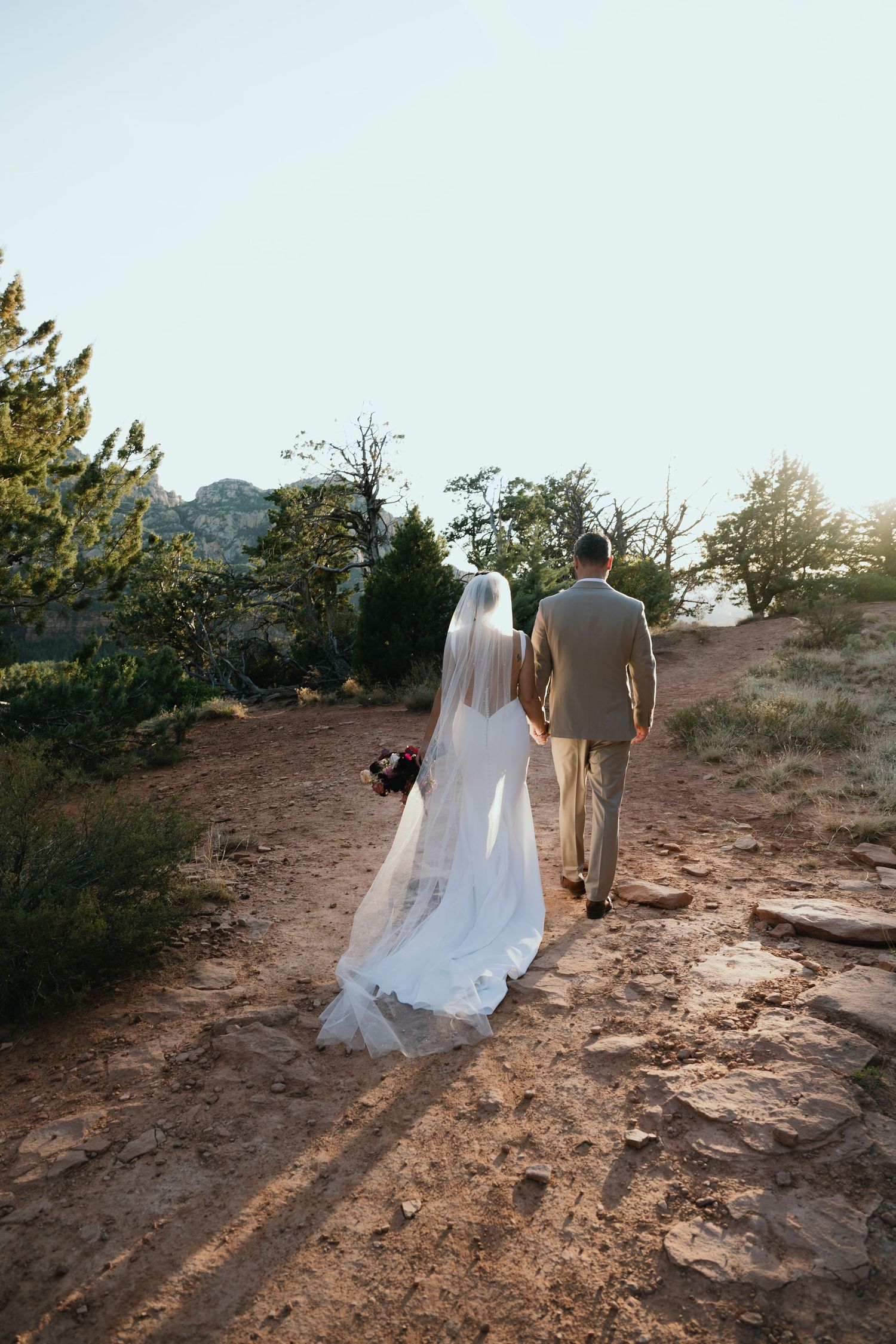 A couple in wedding attire walk along a dusty mountain trail at sunset, the bride's long veil trailing behind them.