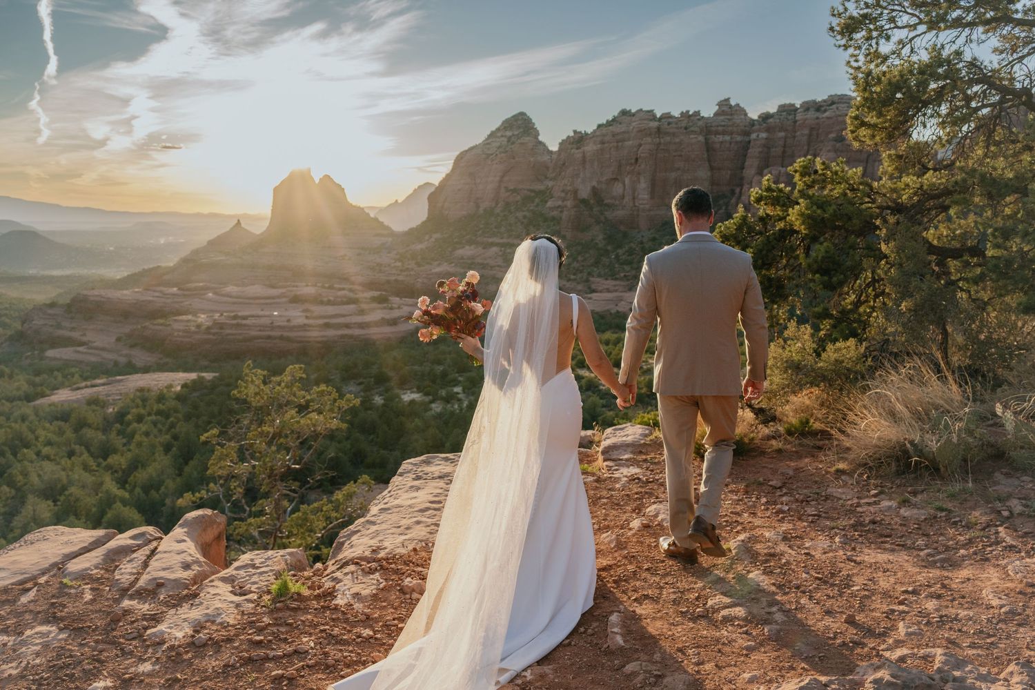 Two wedding day figures stand on a mountain ridge at sunset overlooking red rock formations in a desert landscape.
