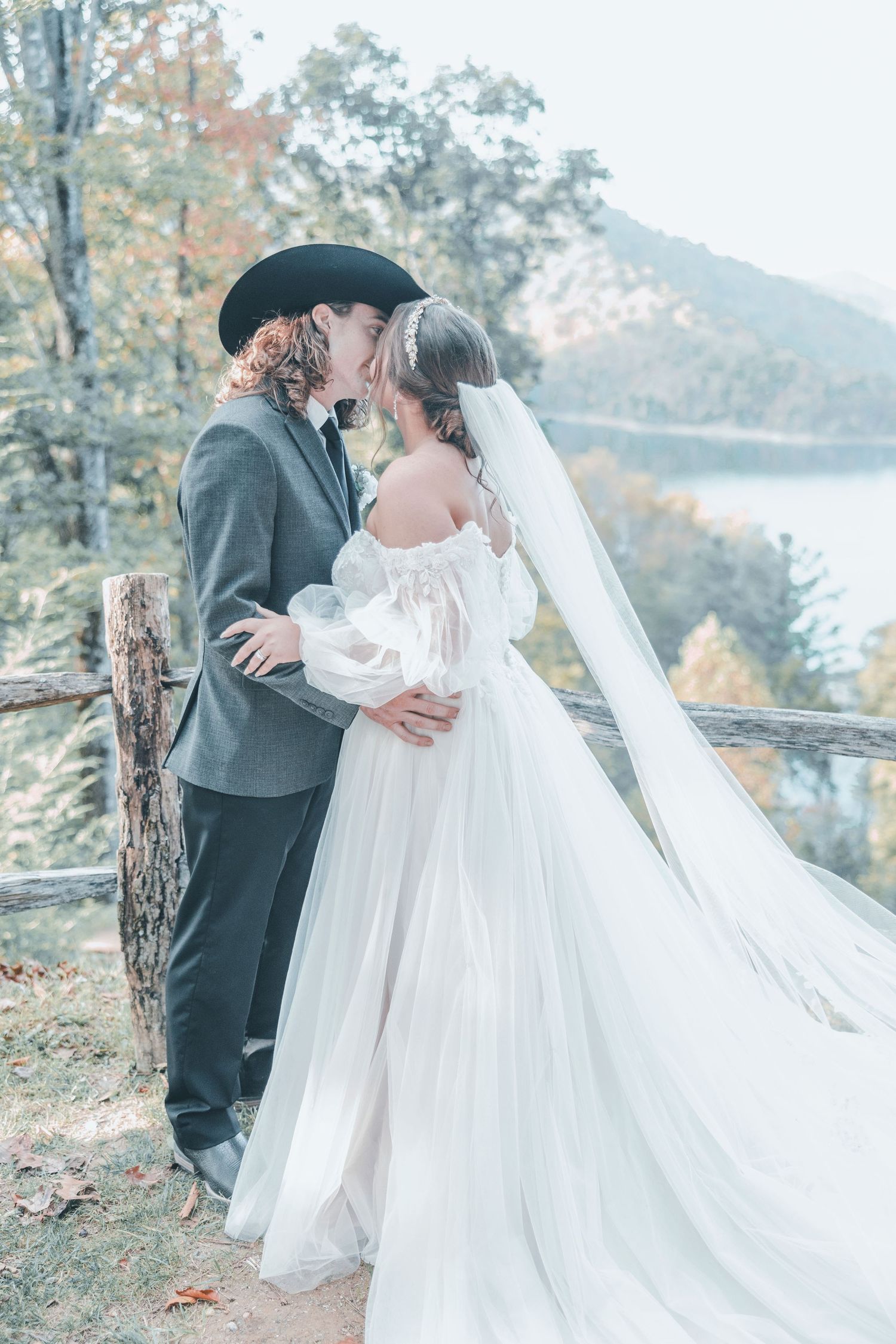 A romantic outdoor wedding scene with a bride in a flowing white dress and groom in grey against an autumn mountain backdrop.