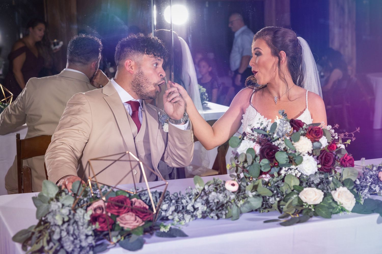 Newlyweds share a candid moment at reception table decorated with lush greenery and floral centerpieces.