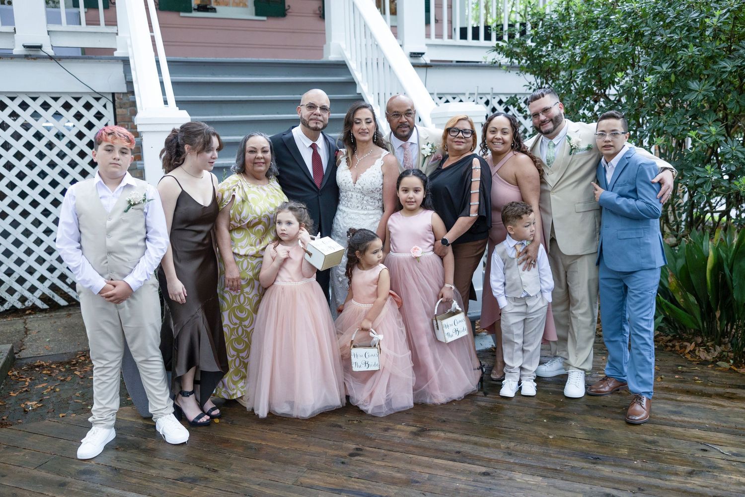 Wedding party poses together on wooden deck outside house wearing pink, beige and blue formal attire.