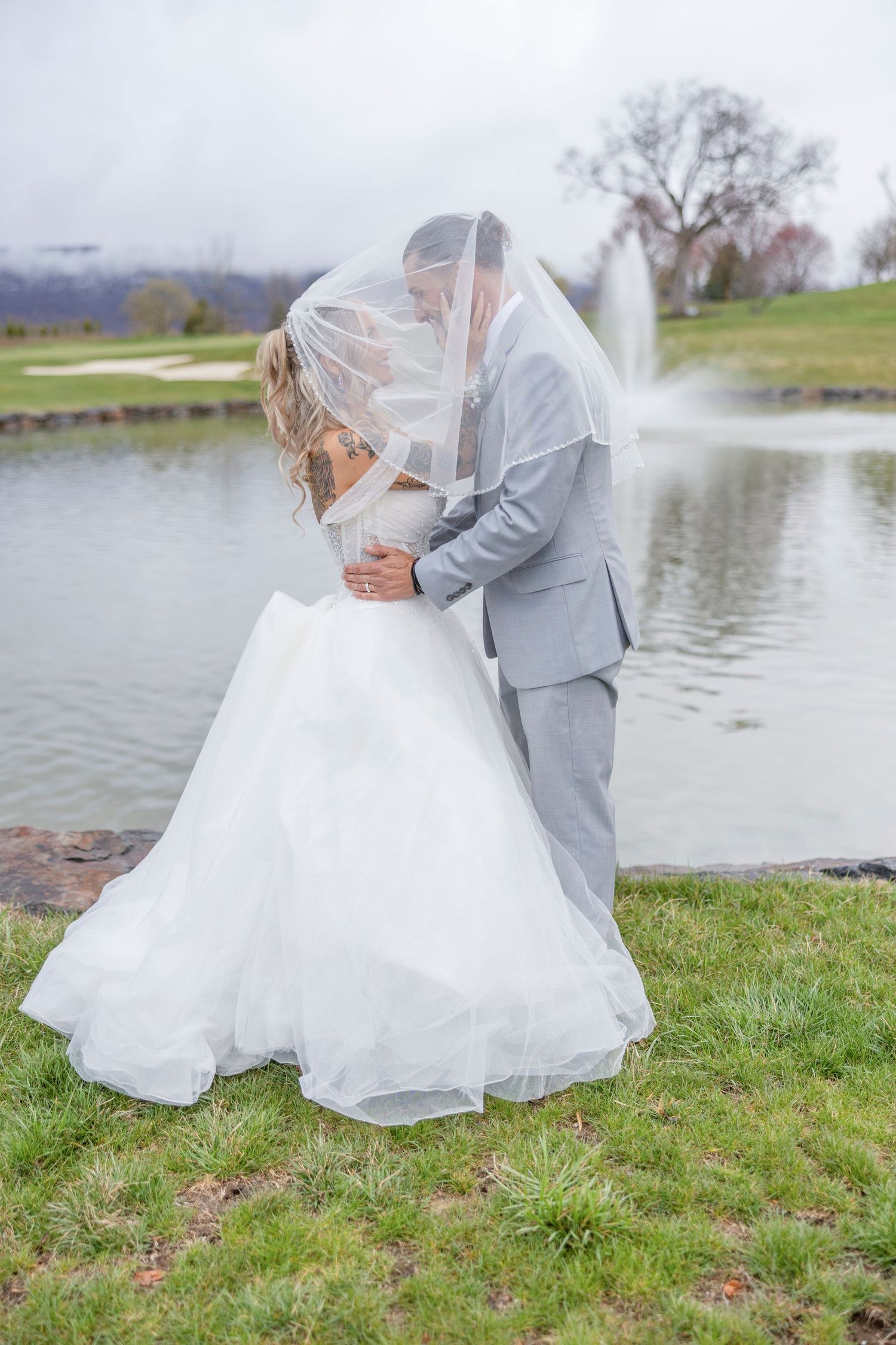 Newlyweds share romantic kiss under flowing veil beside fountain at golf course wedding venue on cloudy day.