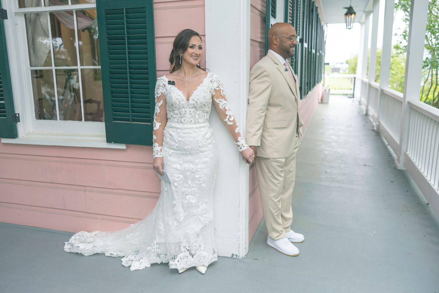 Wedding couple holds hands during their first look moment on a vintage porch with pink walls and green shutters.
