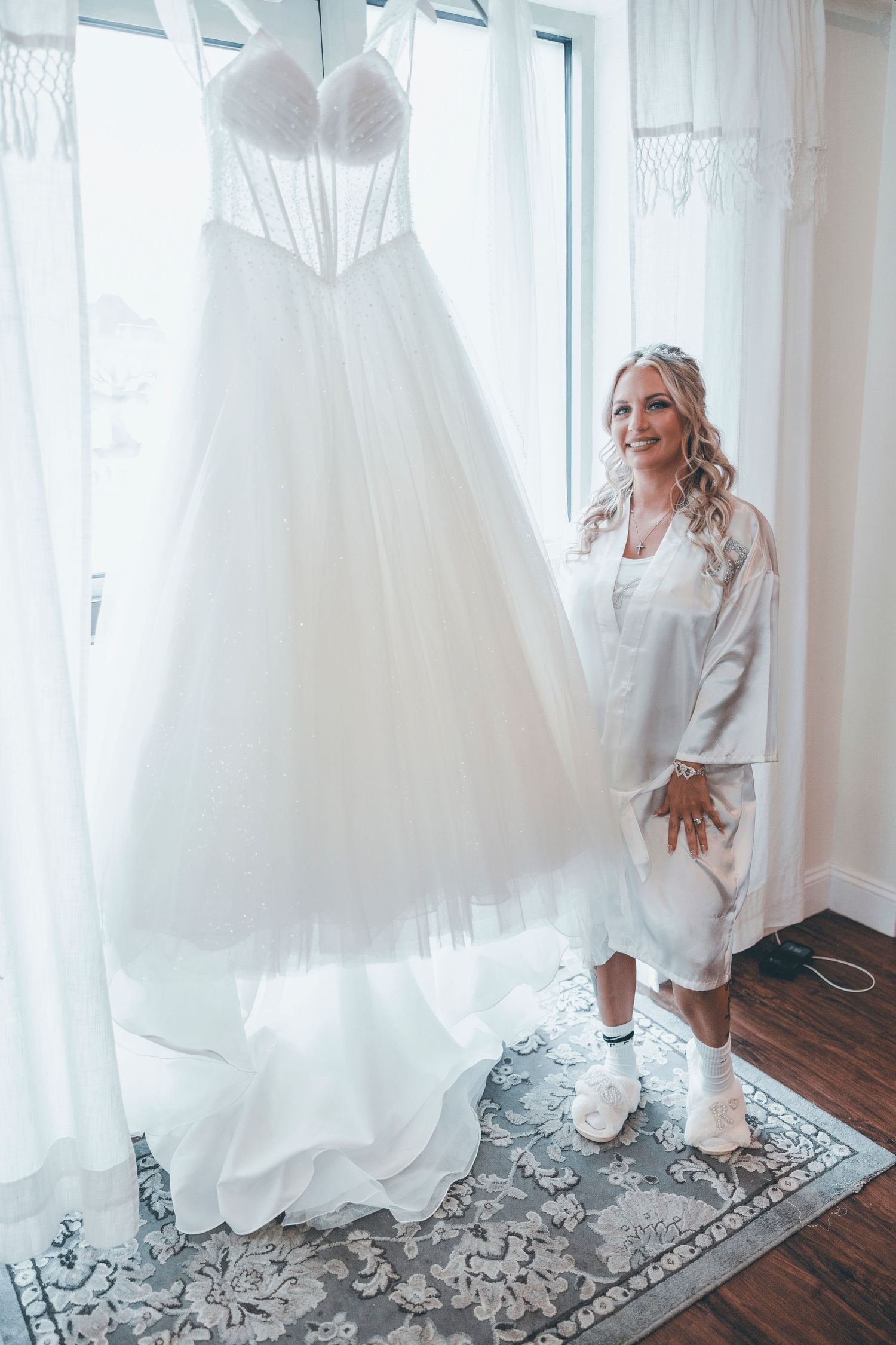 A white strapless wedding dress hangs by a window next to someone wearing a white silk robe on a patterned area rug.