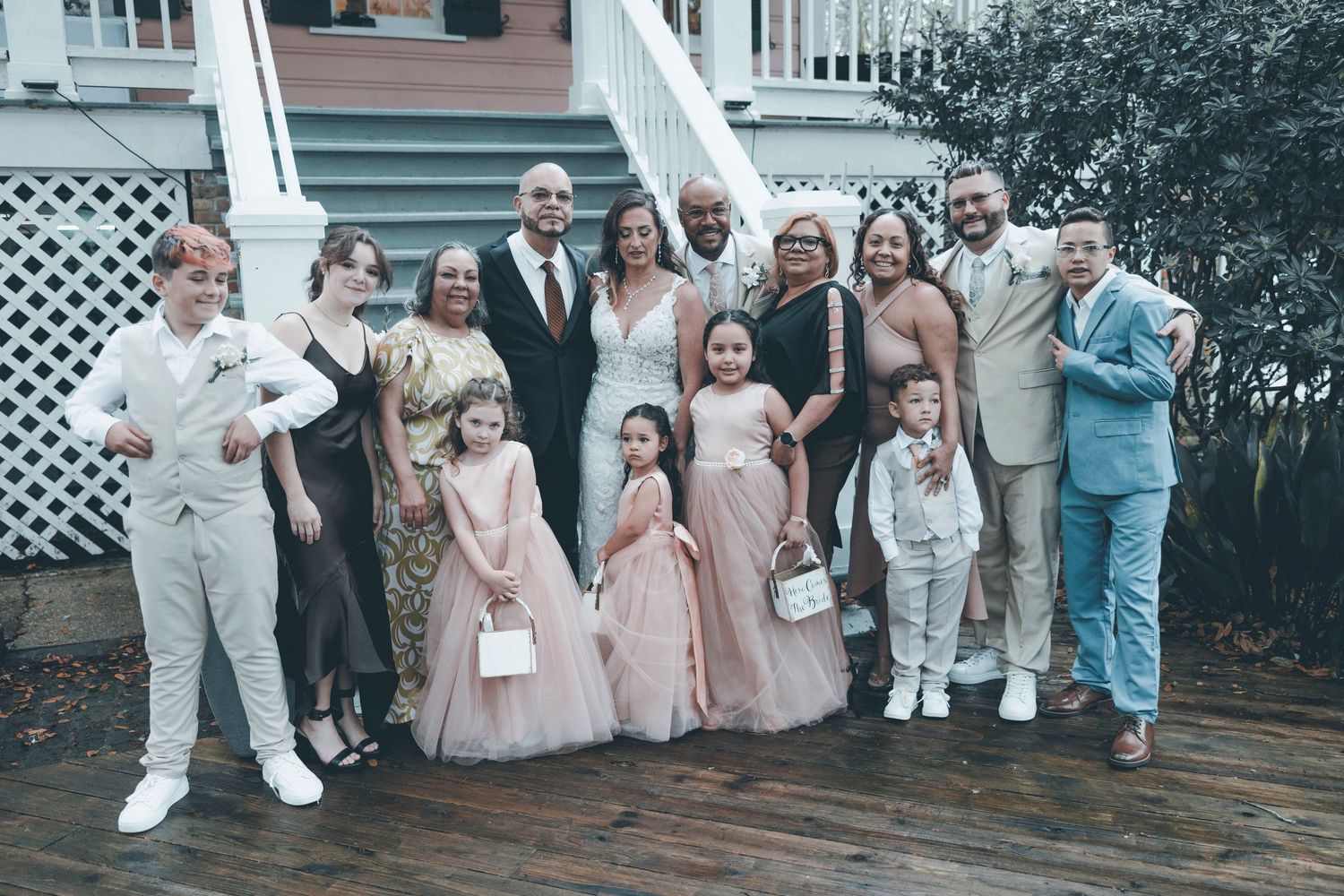 A wedding party in pastel colors and formal attire poses together on wooden deck stairs outside.