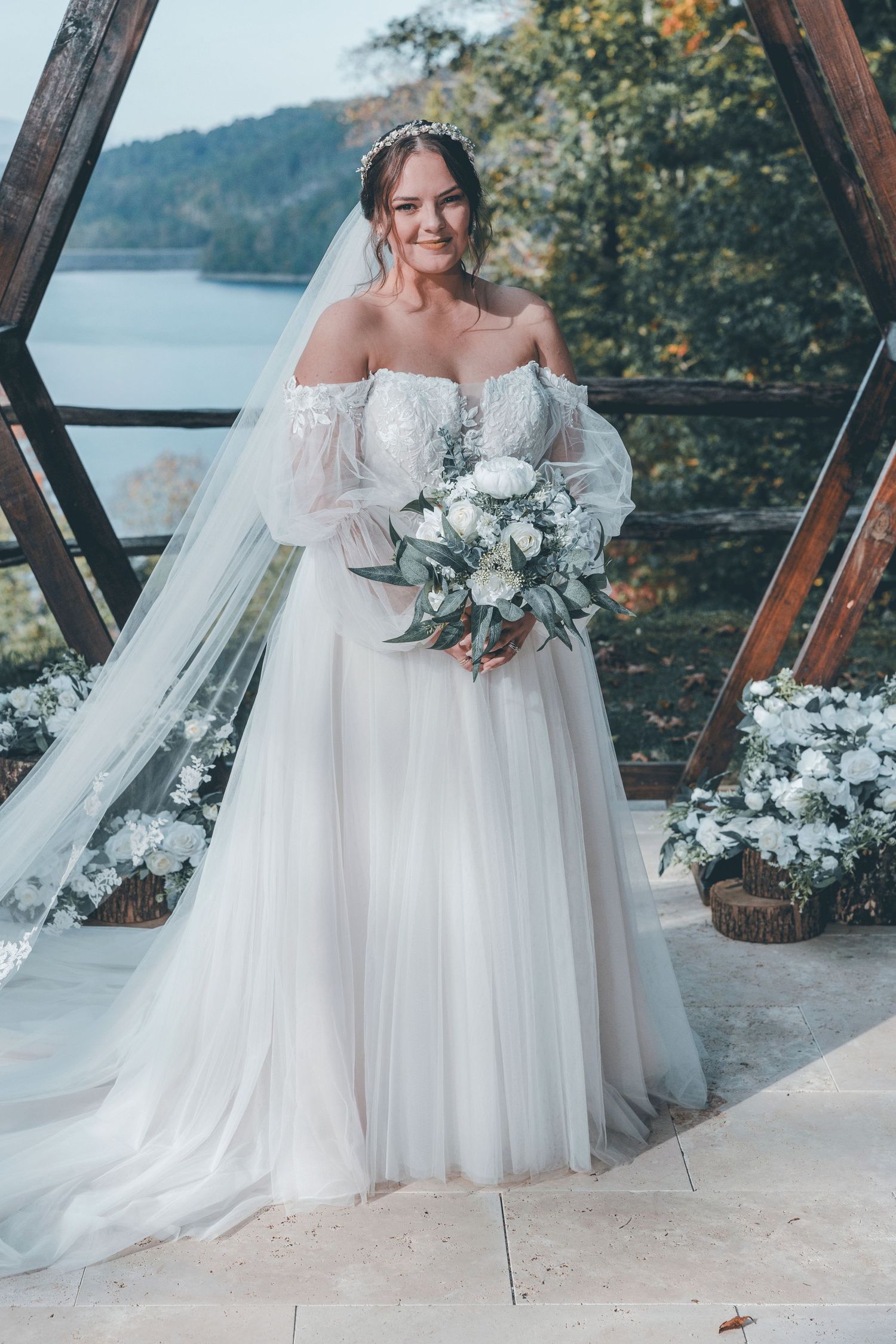 A bride in an ethereal off-shoulder white gown with a flowy veil poses with a eucalyptus bouquet against a lakeside backdrop.