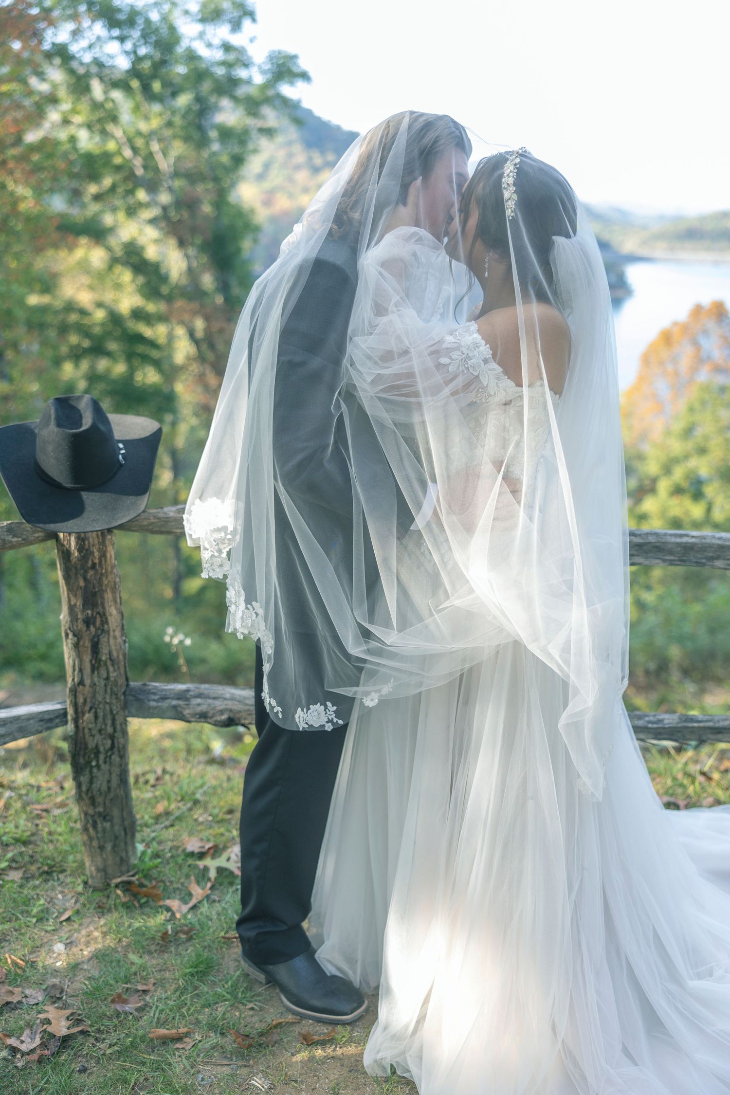 A romantic outdoor wedding moment with two people embracing under flowing white veils beside a scenic lake view.