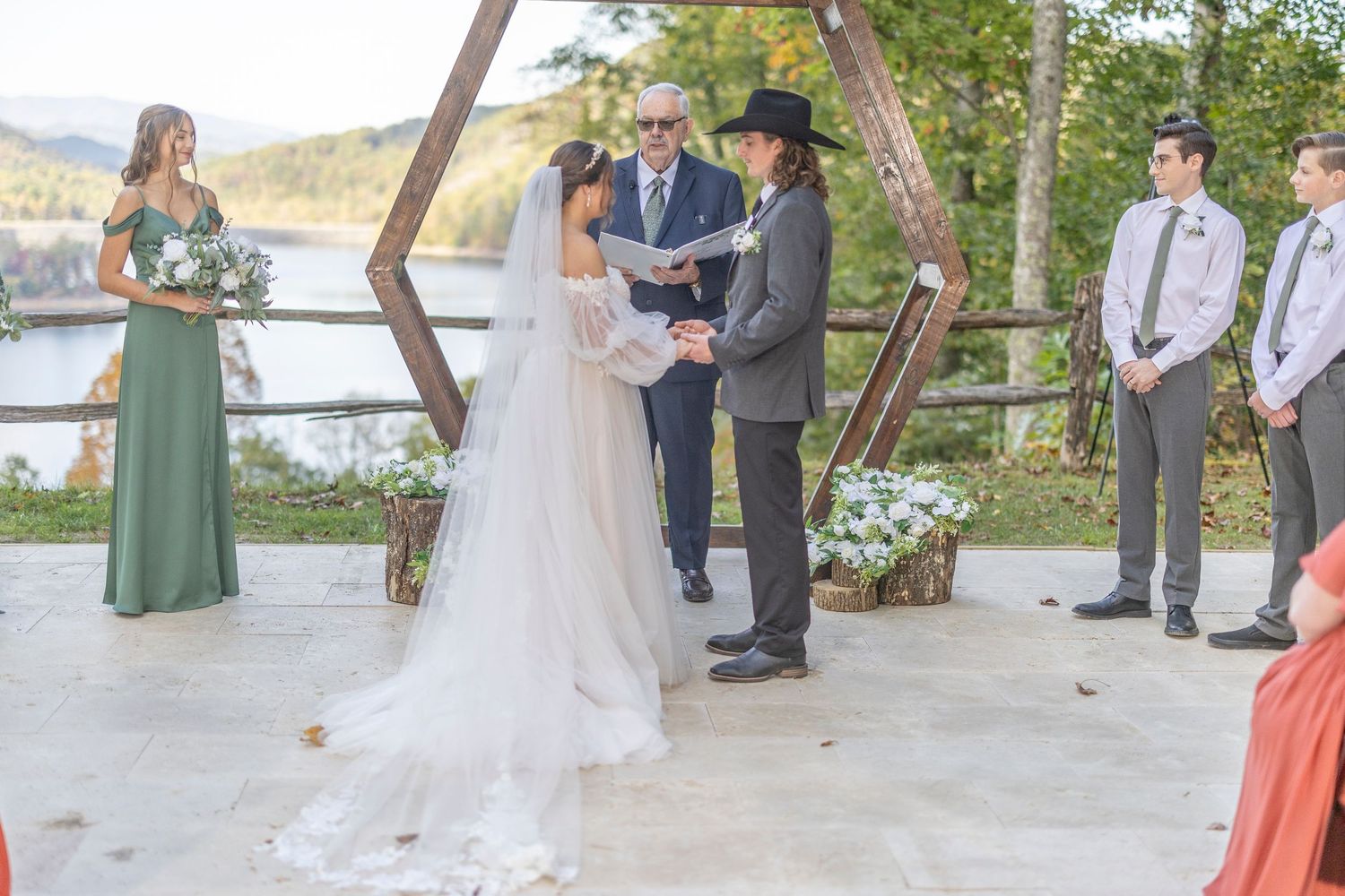 A wedding ceremony taking place outdoors under a wooden hexagonal arch with mountains visible in the background.