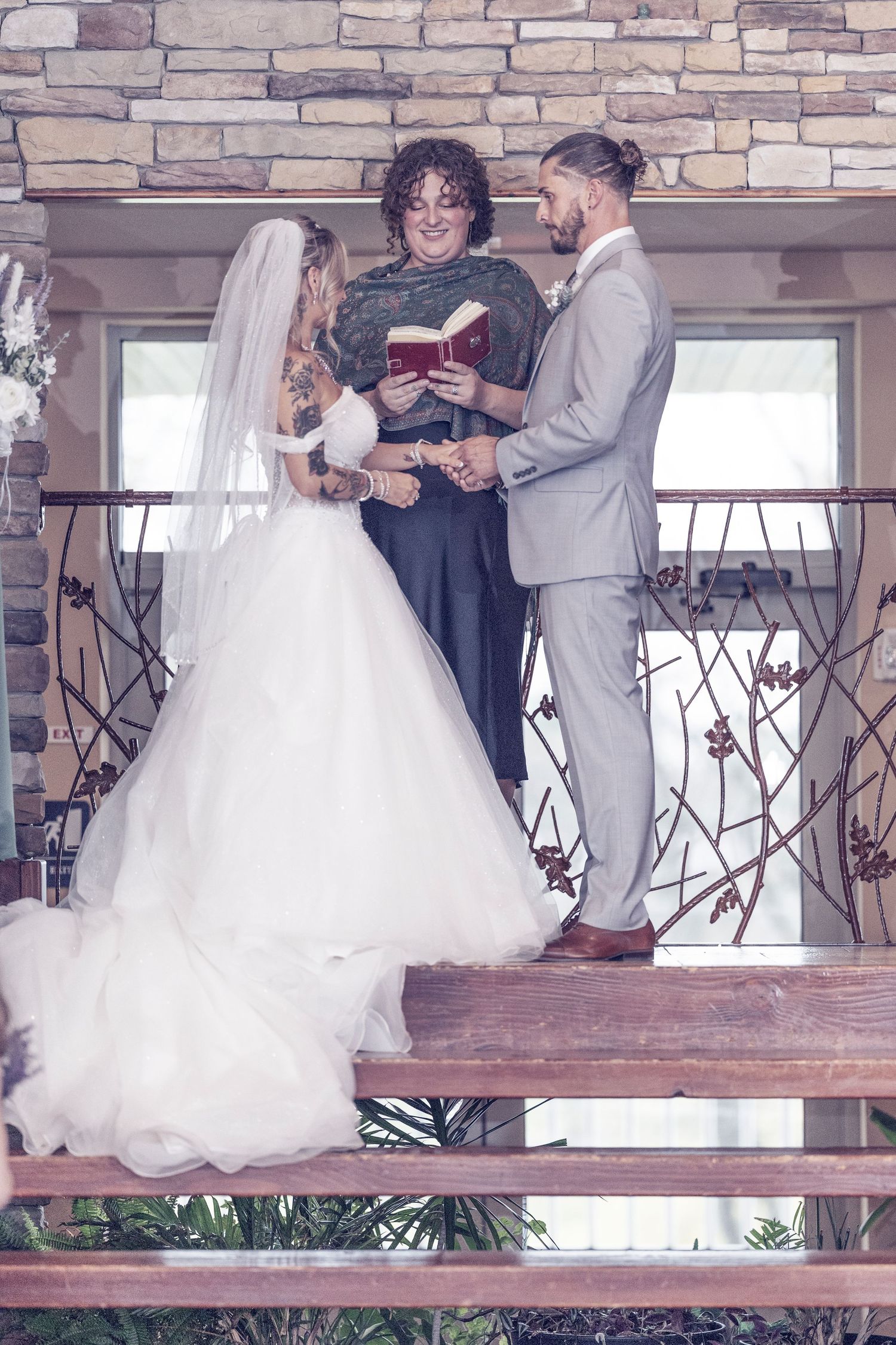 Couple exchanging vows during outdoor wedding ceremony on wooden platform with stone wall backdrop.