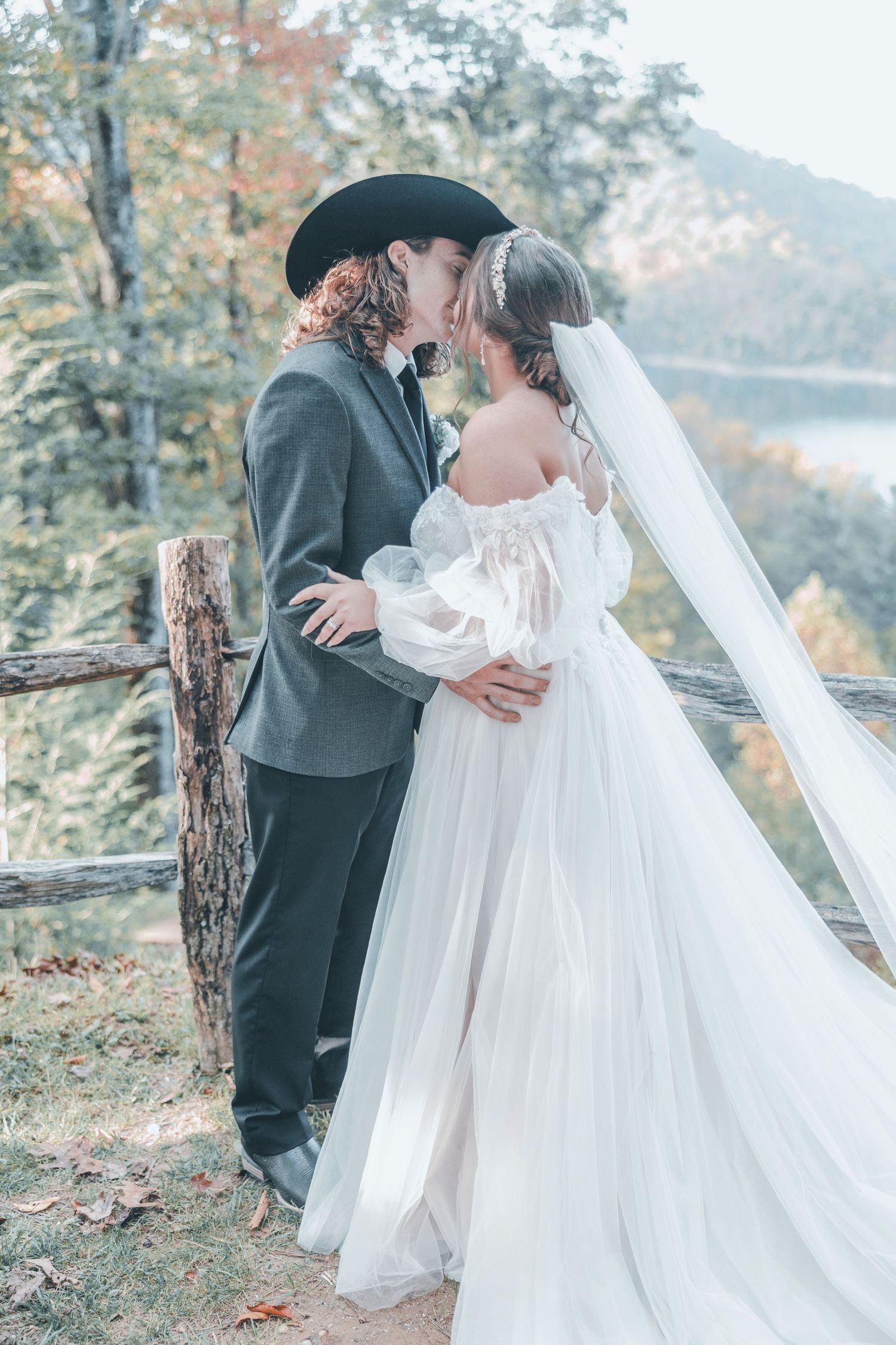 A couple shares a romantic wedding moment against an autumn backdrop with lake and fall foliage in the background.