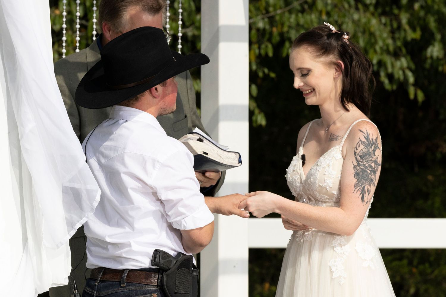 A wedding ceremony exchange of rings taking place under greenery with a bride in white and western-styled groom.