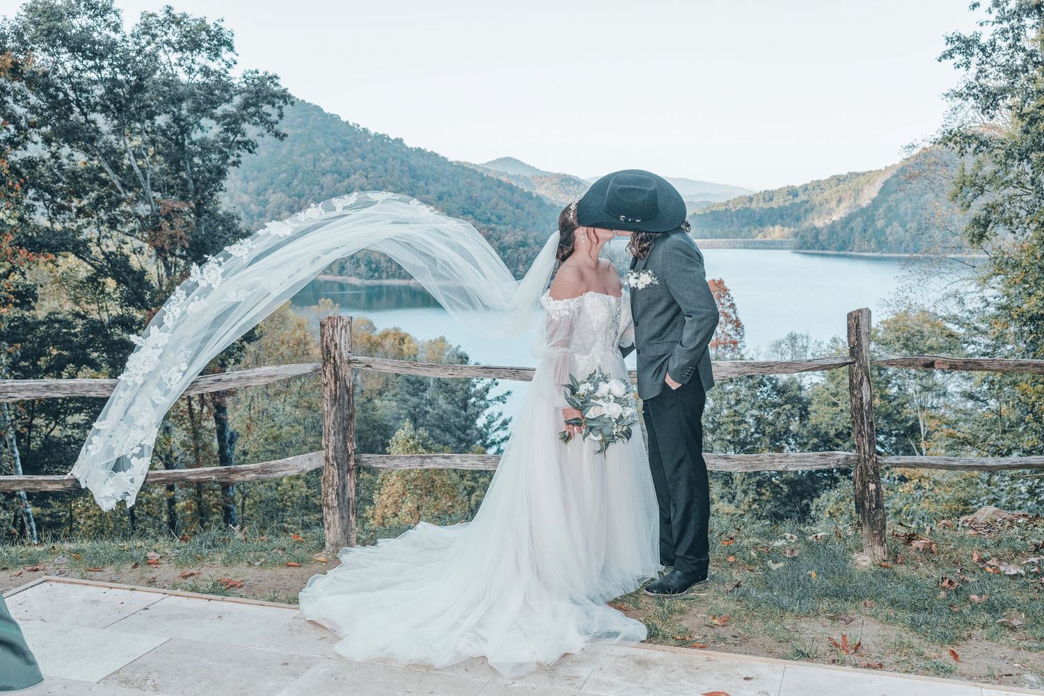 A couple in wedding attire embrace at scenic mountain overlook as long white veil flows dramatically in the breeze.