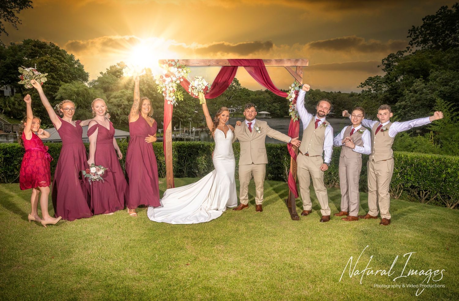 Wedding party celebrates under a pink arch at sunset with bridesmaids in burgundy dresses and groomsmen in tan suits.