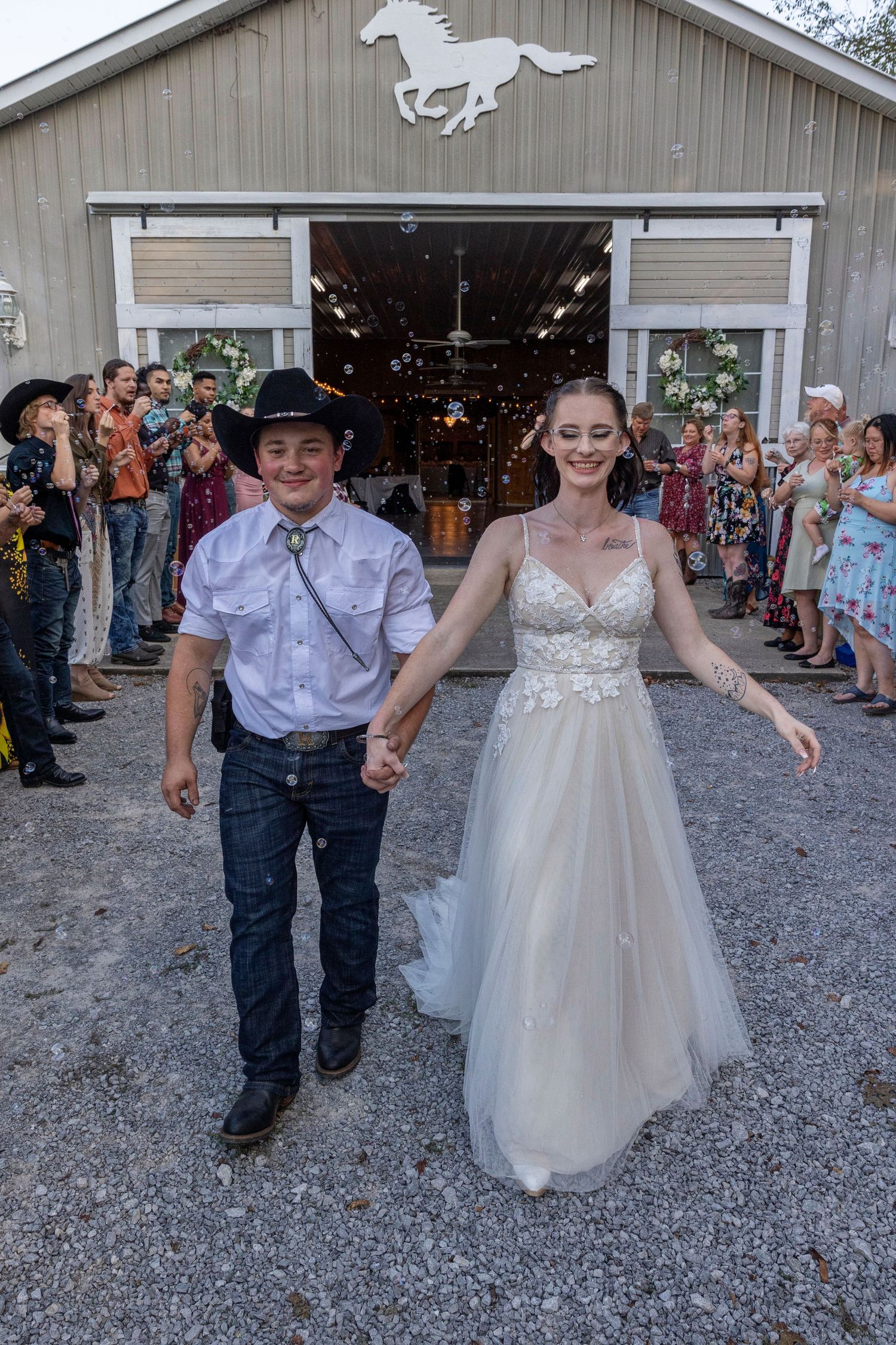 A western-themed wedding couple exits a barn with a horse silhouette, surrounded by guests on a gravel path.
