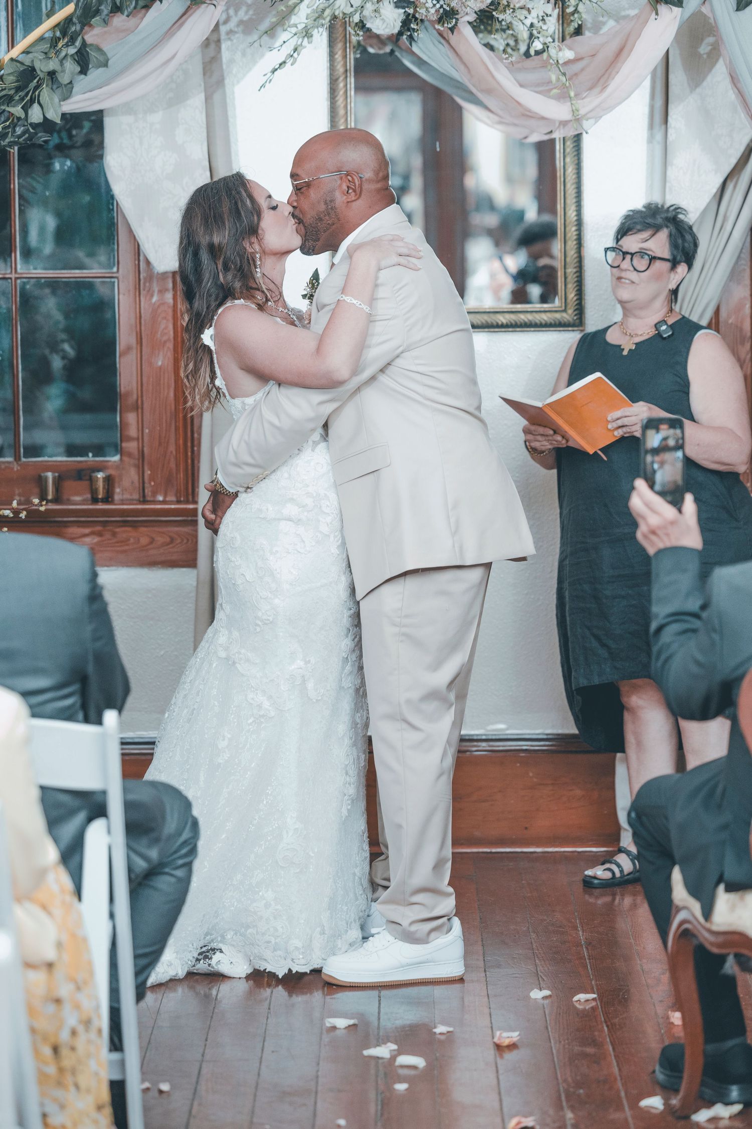 A romantic first kiss at a wedding ceremony as an officiant holds a book during an indoor celebration.