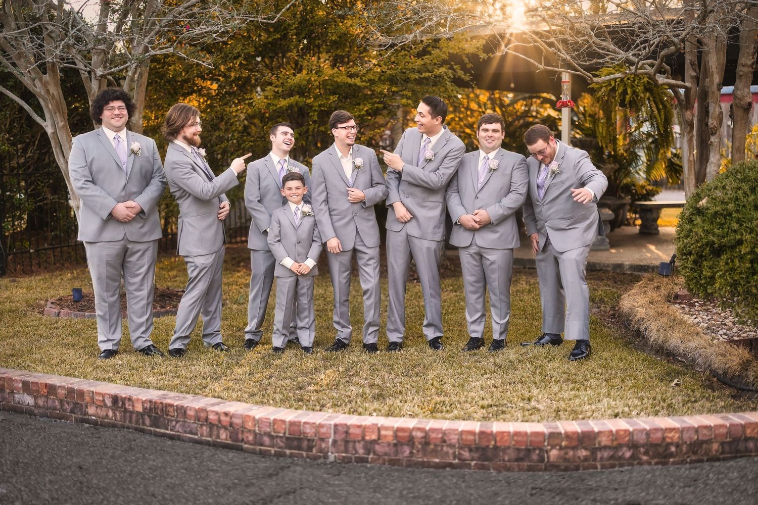A group wearing matching light gray suits poses together outdoors at sunset with trees in the background.