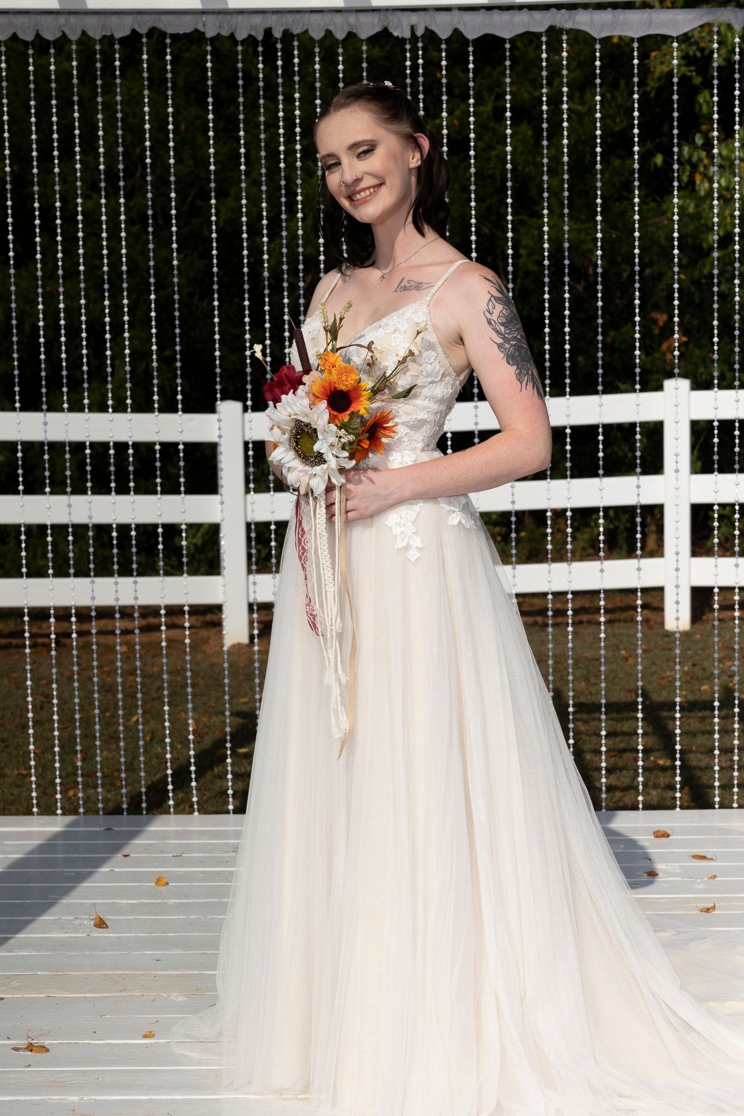 Person in white wedding dress holding autumn bouquet poses on deck with string light backdrop.