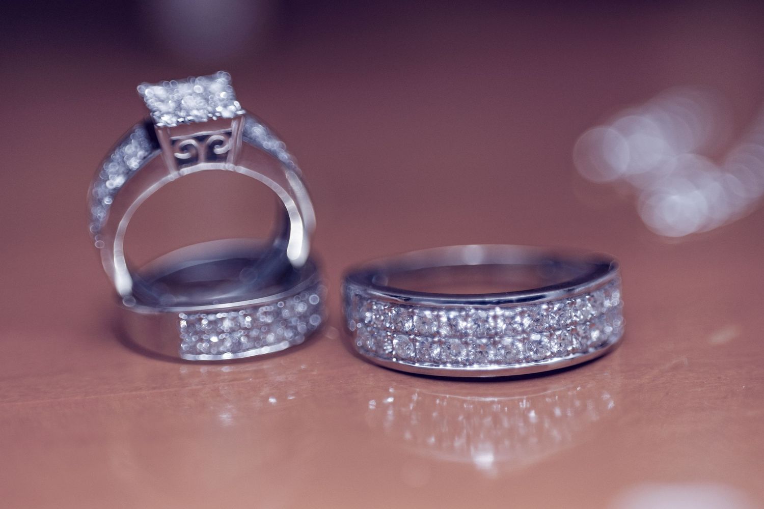 Two diamond wedding rings with elaborate settings photographed on a brown surface with bokeh lights in background.