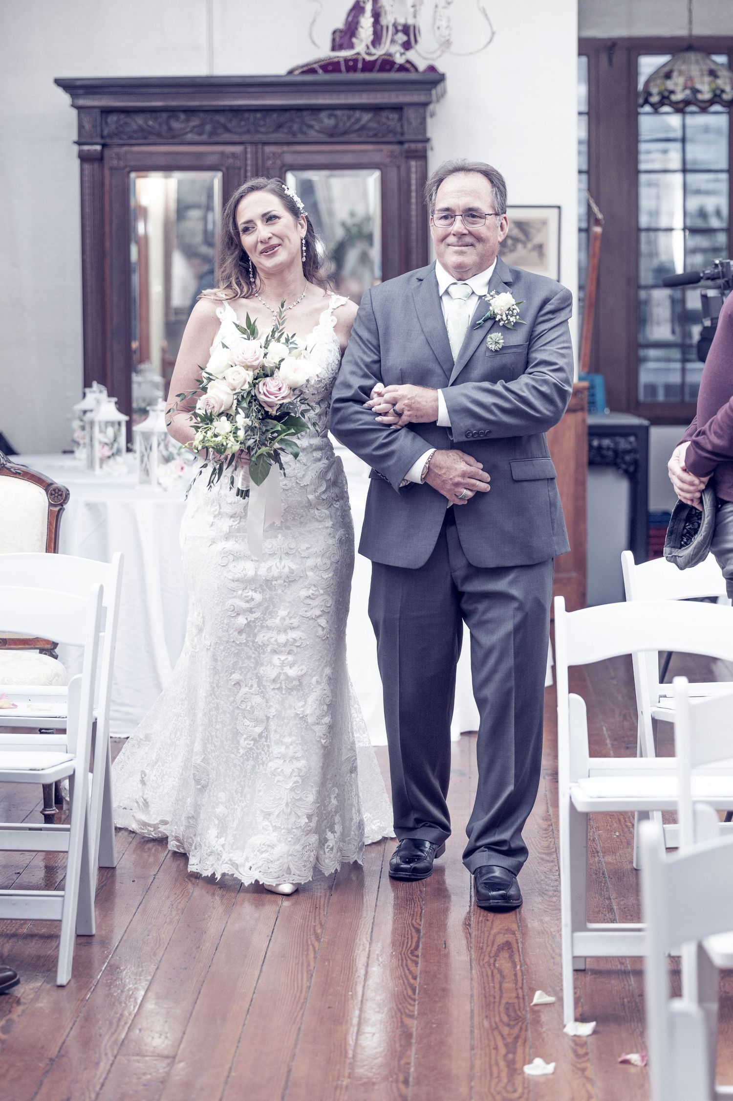 A couple stands together during their wedding ceremony on a wooden deck with white chairs and antique furniture in background.