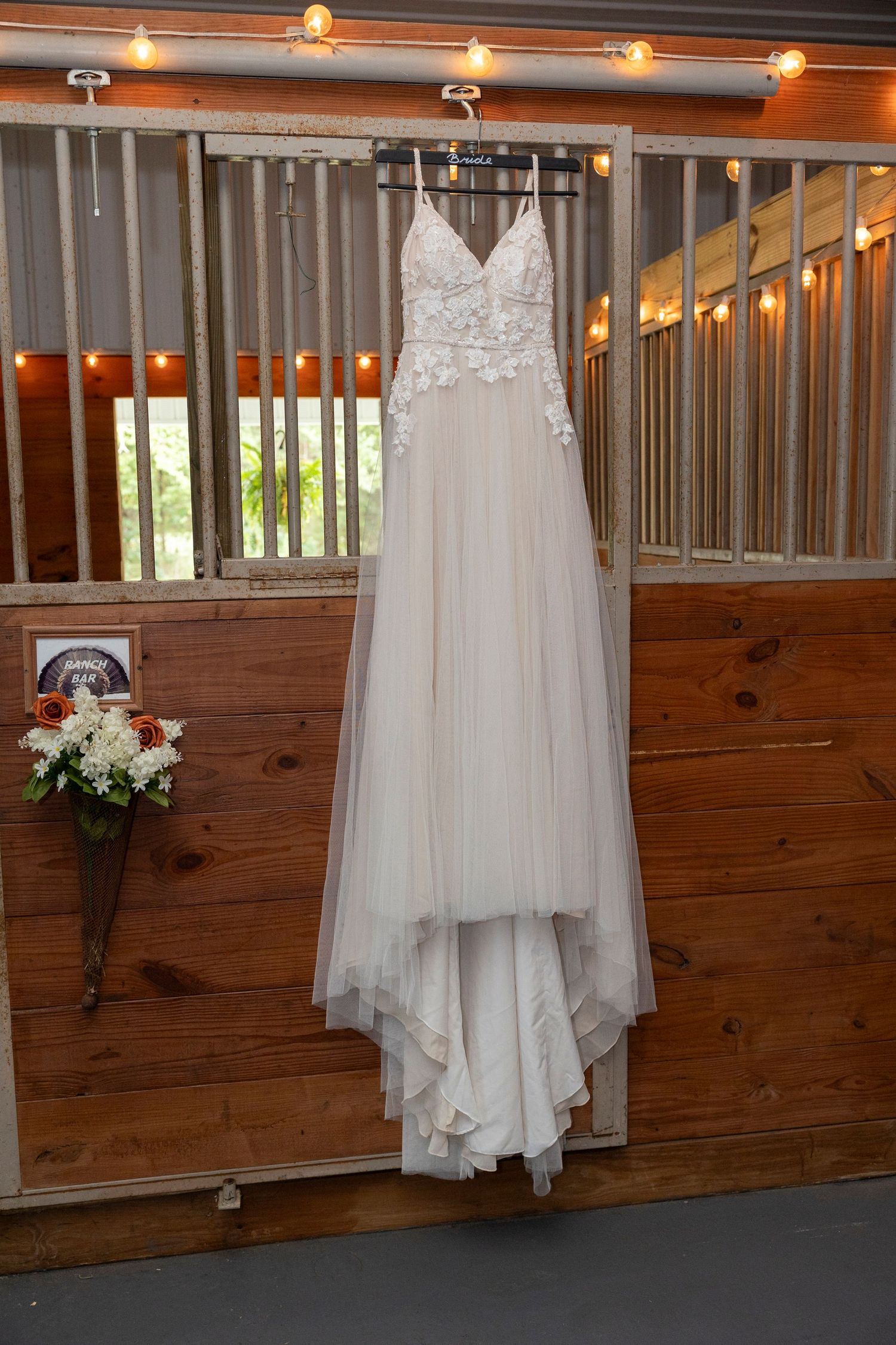 A white lace and tulle wedding dress hangs elegantly against a rustic wooden wall with warm overhead lighting.