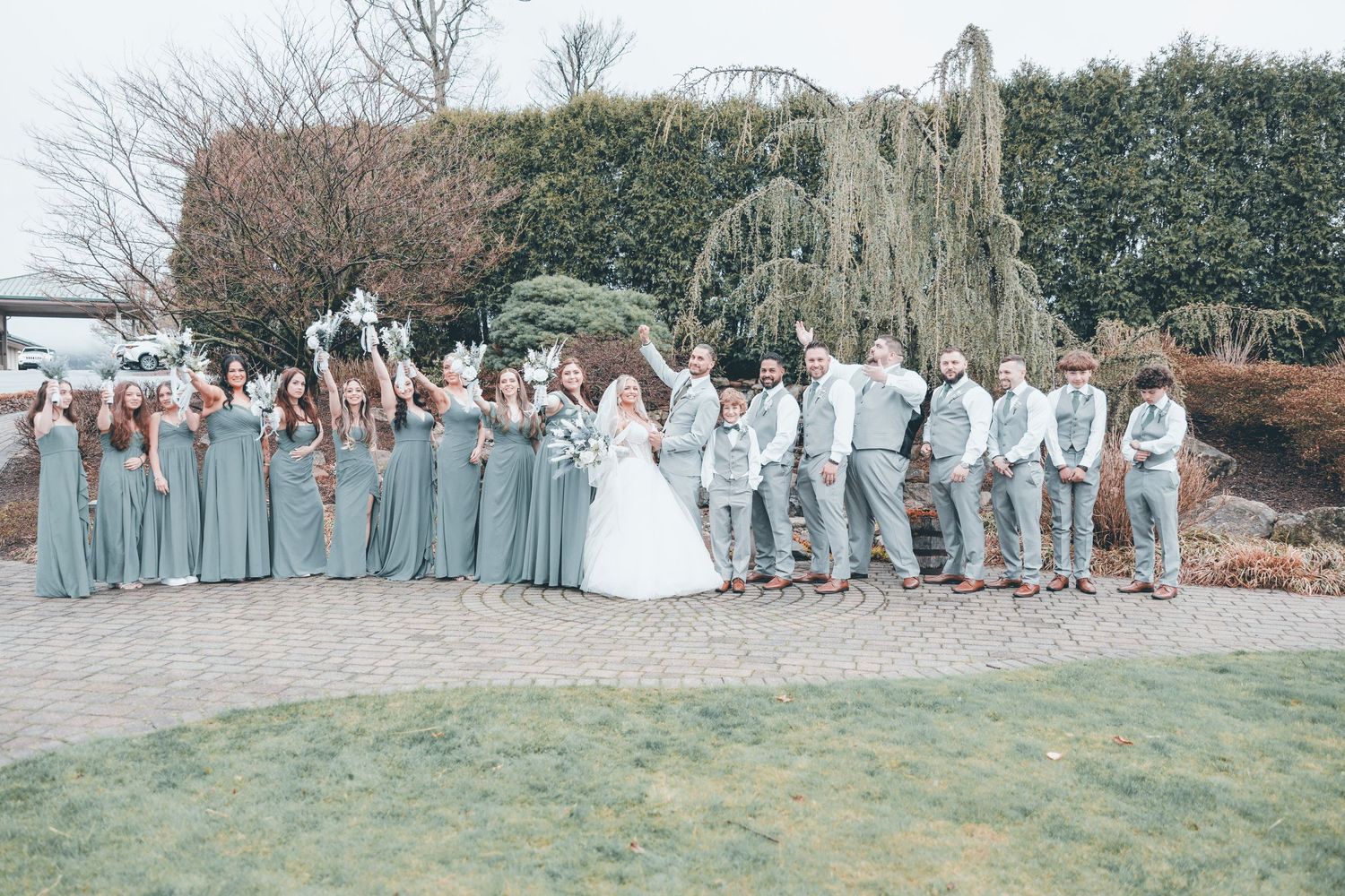 Wedding party in light blue bridesmaid dresses and gray suits celebrates joyfully on a paved garden path.