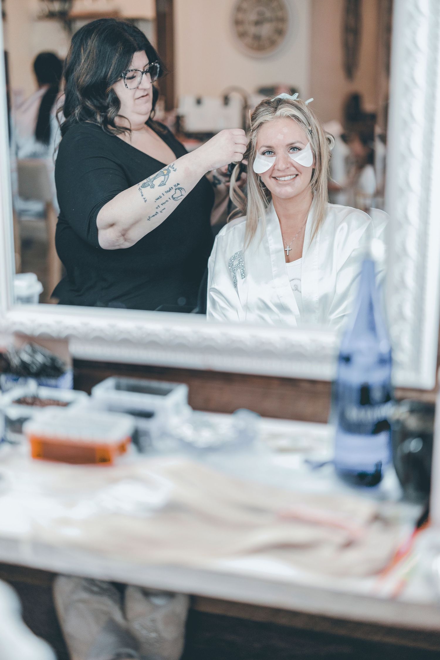 A hairstylist wearing black styles and prepares a client in a white robe at a salon vanity table.