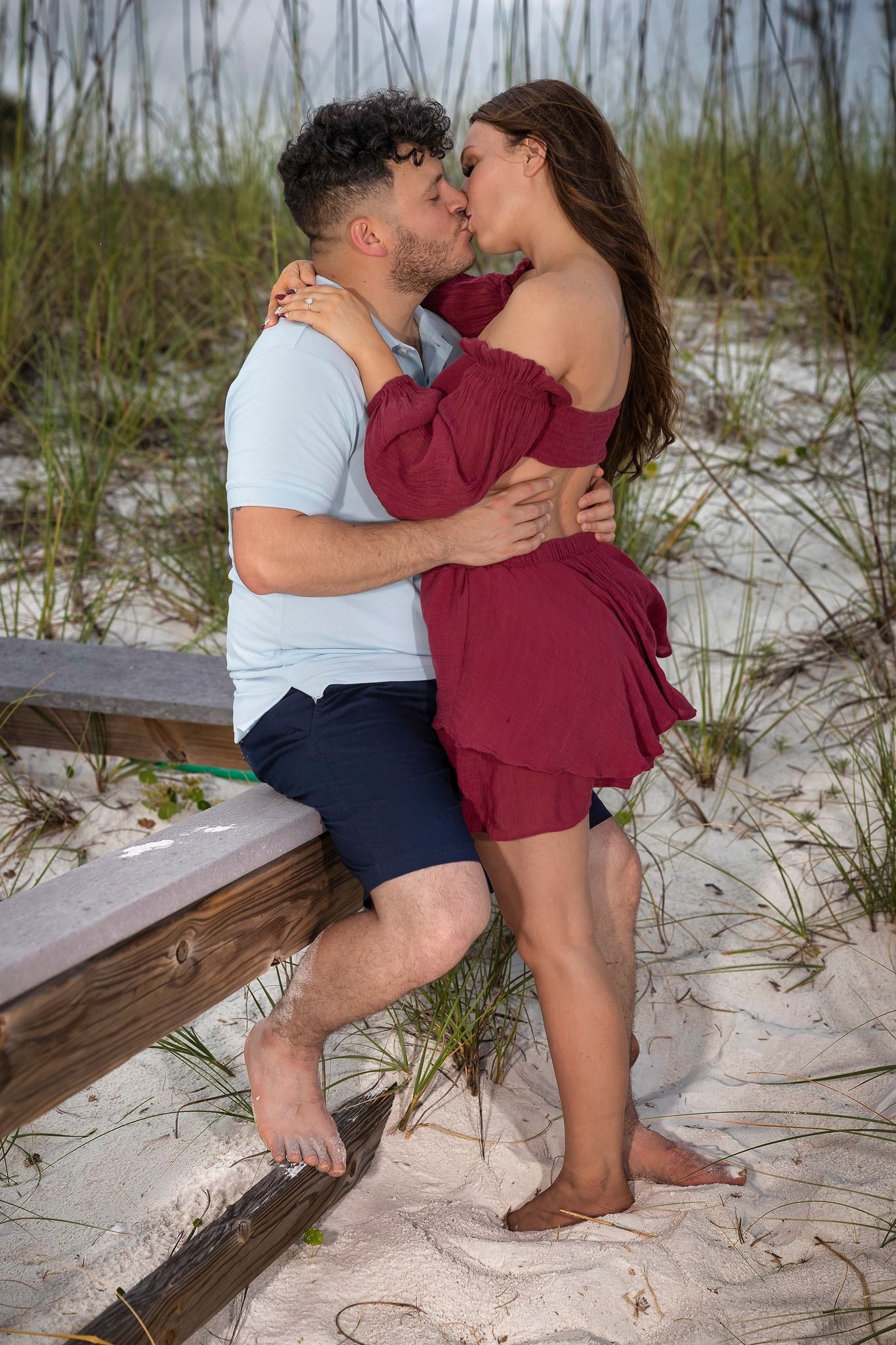 A couple embraces romantically on sandy beach steps with tall grass in the background at dusk.