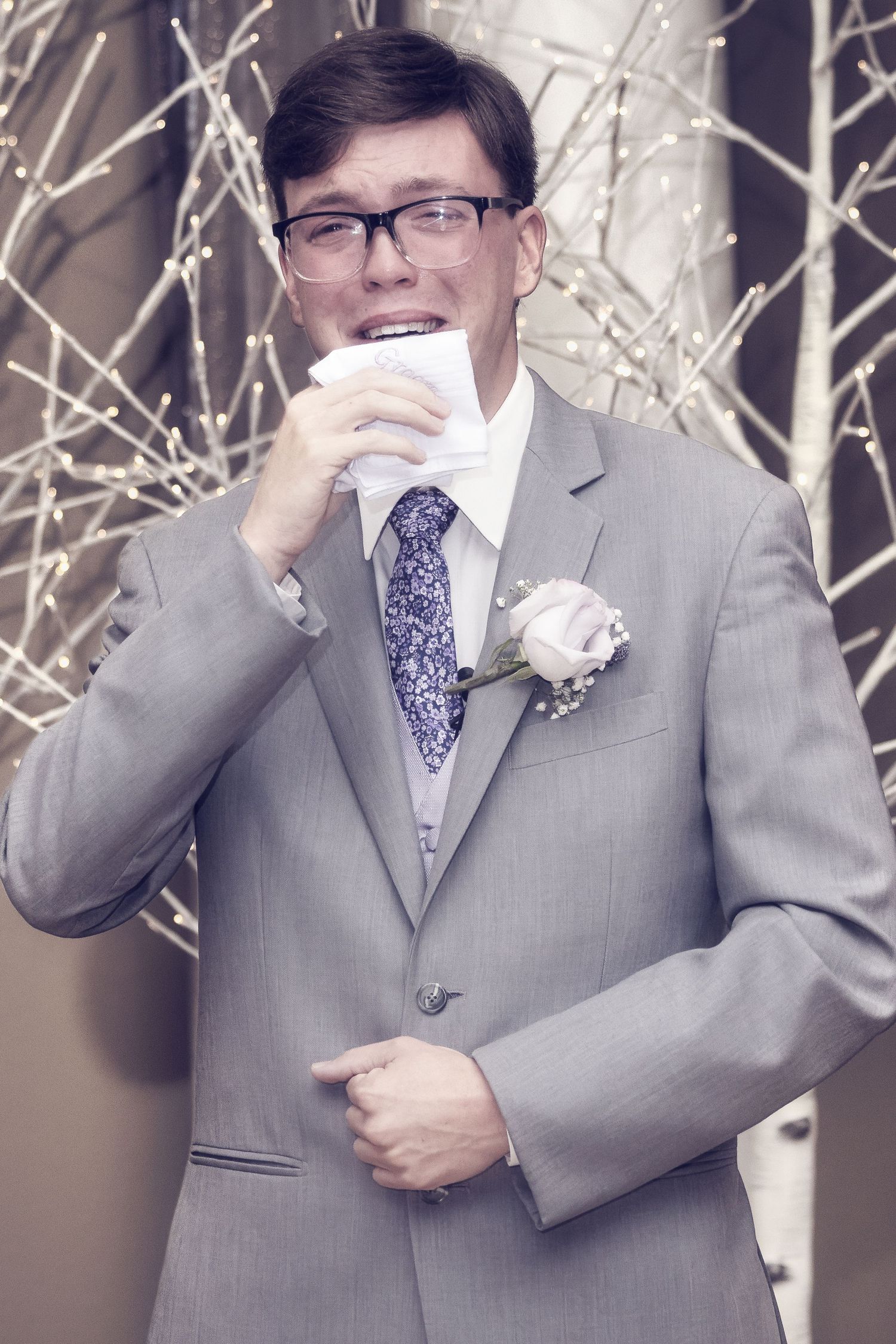Wedding party member in gray suit and tie dabs tears with handkerchief during emotional ceremony moment.