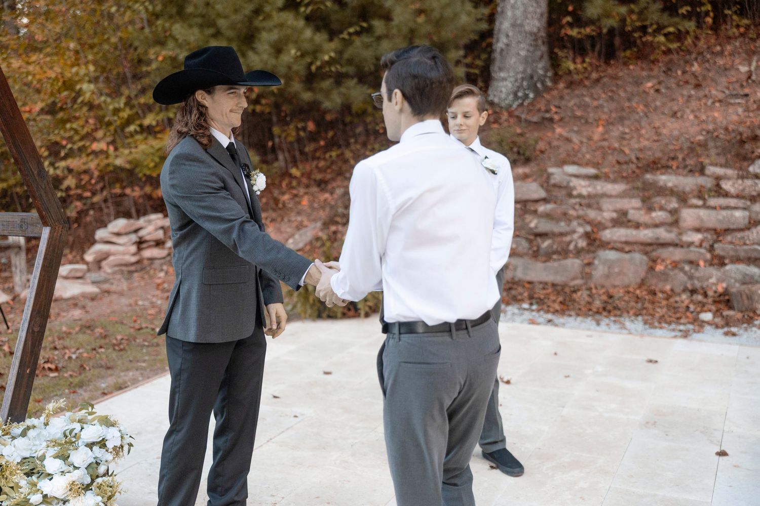 Person in formal attire and cowboy hat shakes hands with another person wearing white shirt on stone path outdoors.