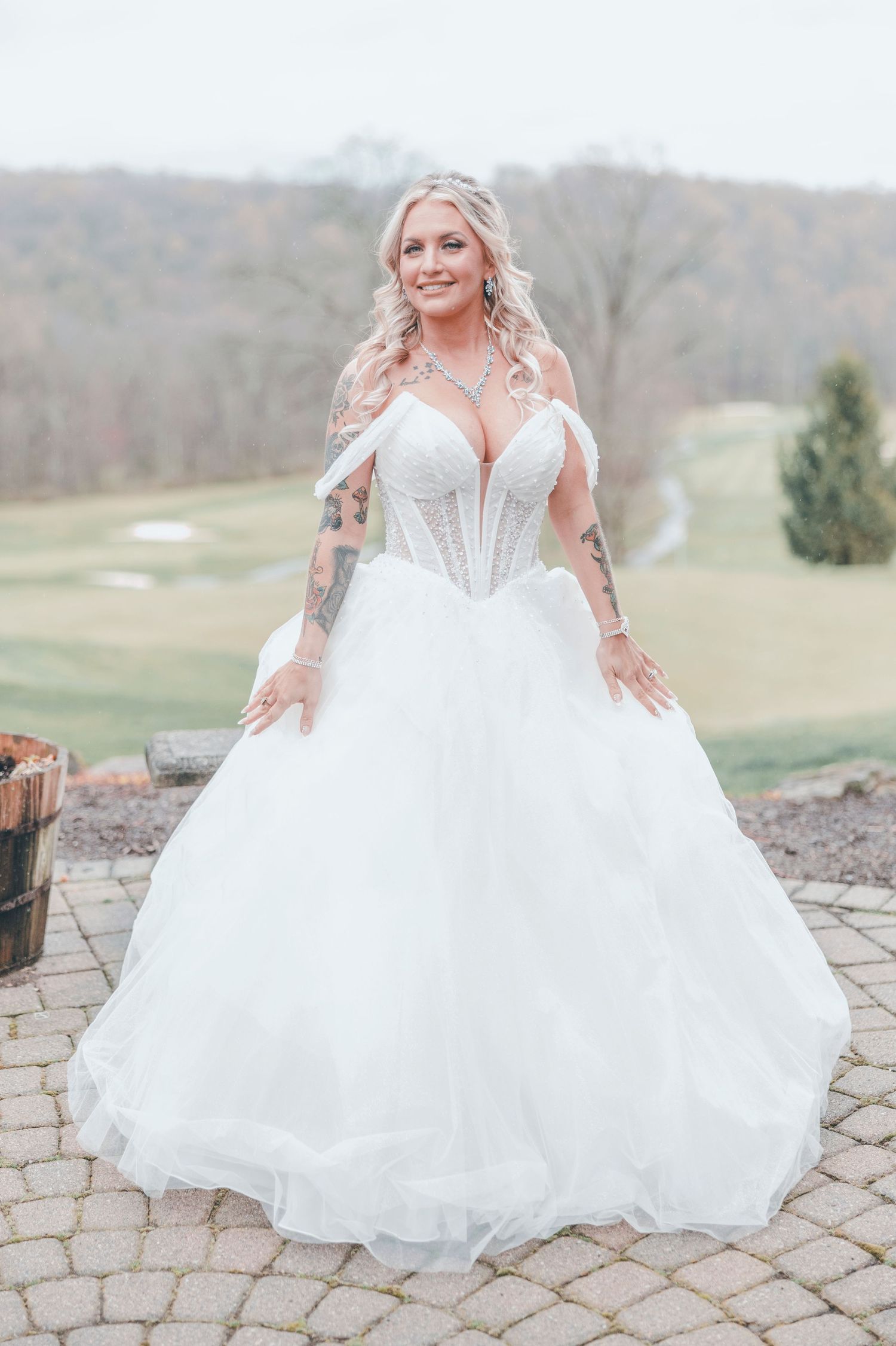 A bride in a stunning white ball gown with tattoos stands on stone pavers with a scenic backdrop.