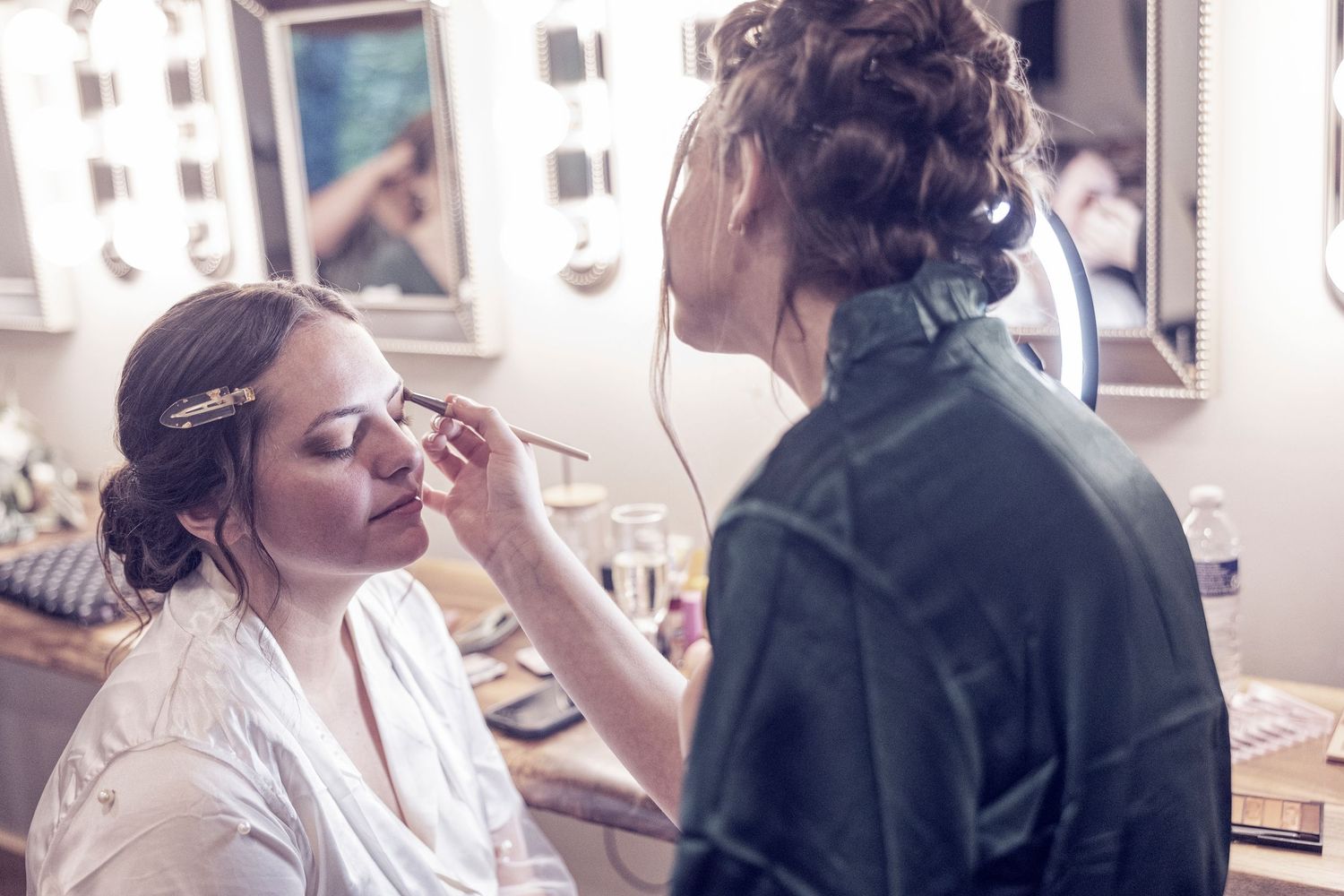 Makeup artist applies eye makeup to woman in white robe at a beauty salon vanity mirror.
